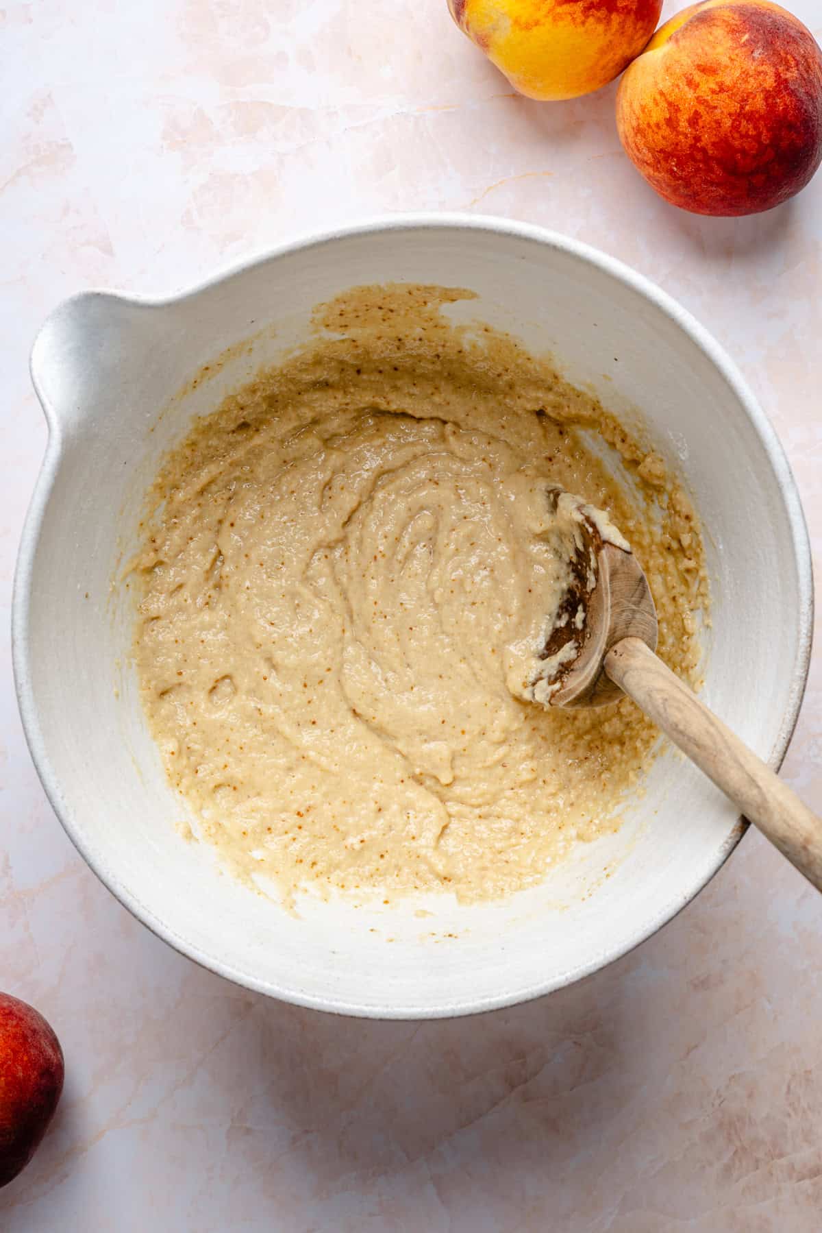 A mixing bowl with thick almond flour peach cobbler batter and a wooden spoon rests on a light surface. Three peaches are nearby.