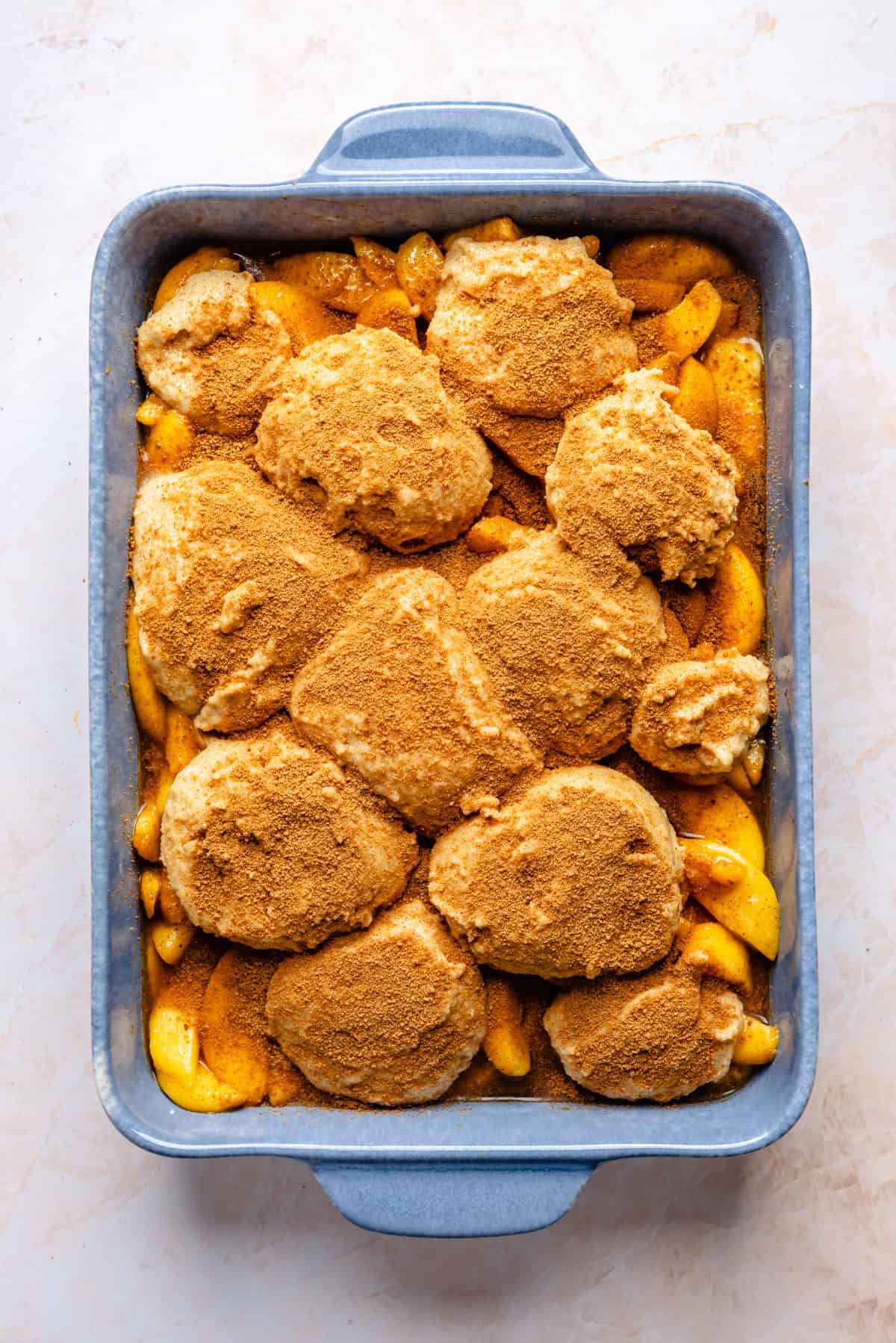 A baking dish filled with sliced peaches topped with gluten free almond flour biscuit dough and a layer of cinnamon, ready to be baked into a delicious gluten free peach cobbler.