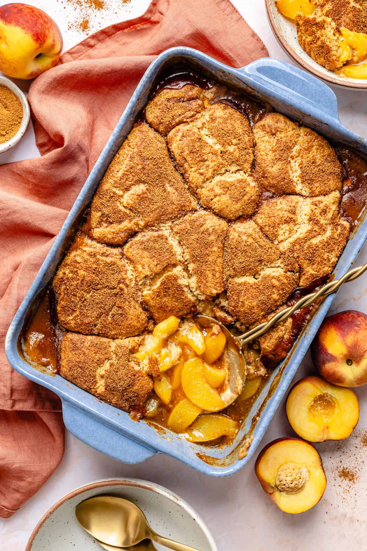 A blue baking dish filled with golden-brown gluten free peach cobbler, with a portion scooped out, surrounded by fresh peaches, a bowl of sliced peaches, a bowl of cinnamon, a napkin, and two gold spoons on a white surface.