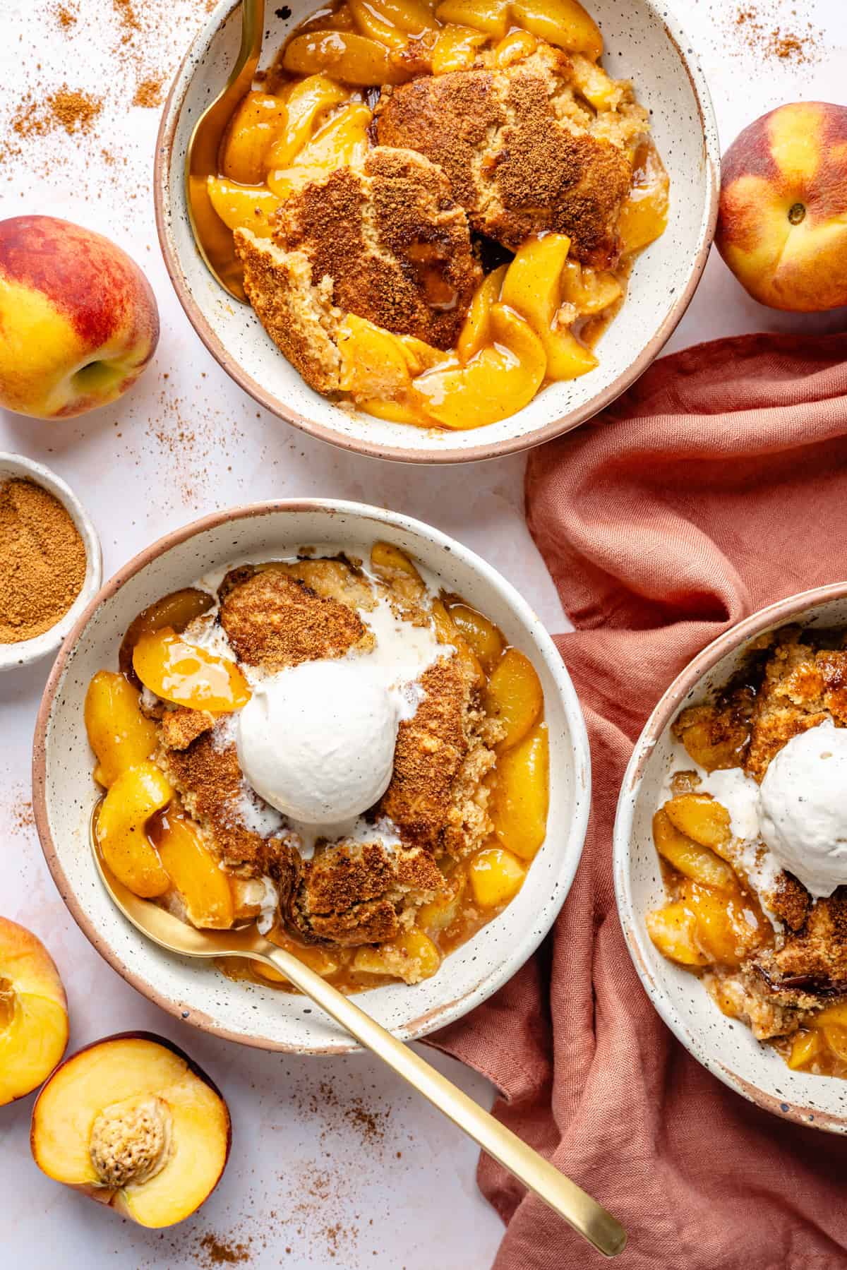 Three bowls of gluten free almond flour peach cobbler, one topped with vanilla ice cream, surrounded by fresh peaches, cinnamon, and a rust-colored napkin on a white surface.