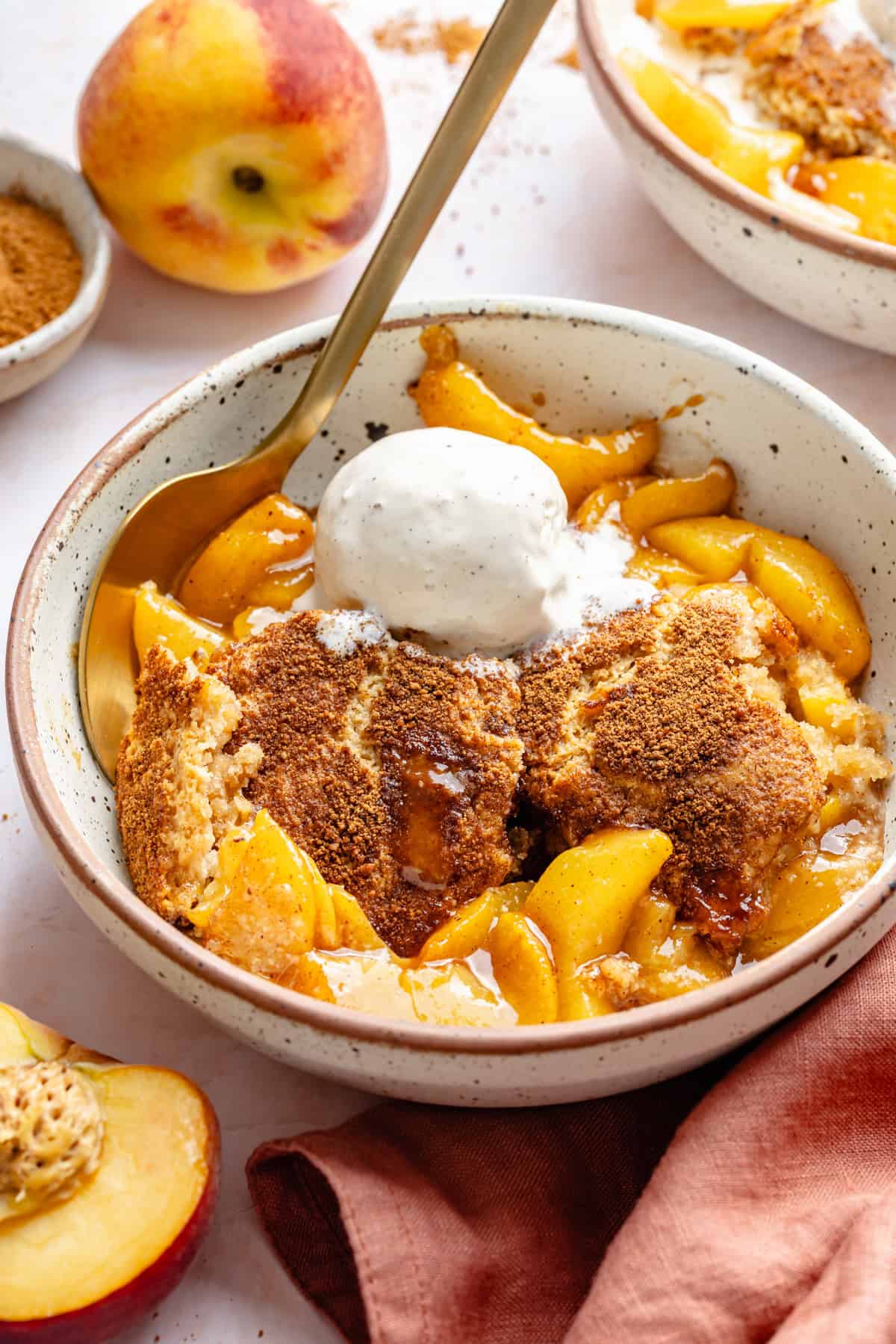 A bowl of almond flour peach cobbler topped with vanilla ice cream, featuring a golden baked crust, juicy peach slices, and a spoon. Fresh peaches and a brown napkin are nearby.