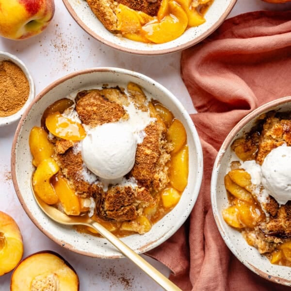 Bowls of gluten free almond flour peach cobbler topped with a scoop of vanilla ice cream, surrounded by fresh peaches, a jar of brown sugar, and a rust-colored napkin on a white surface.