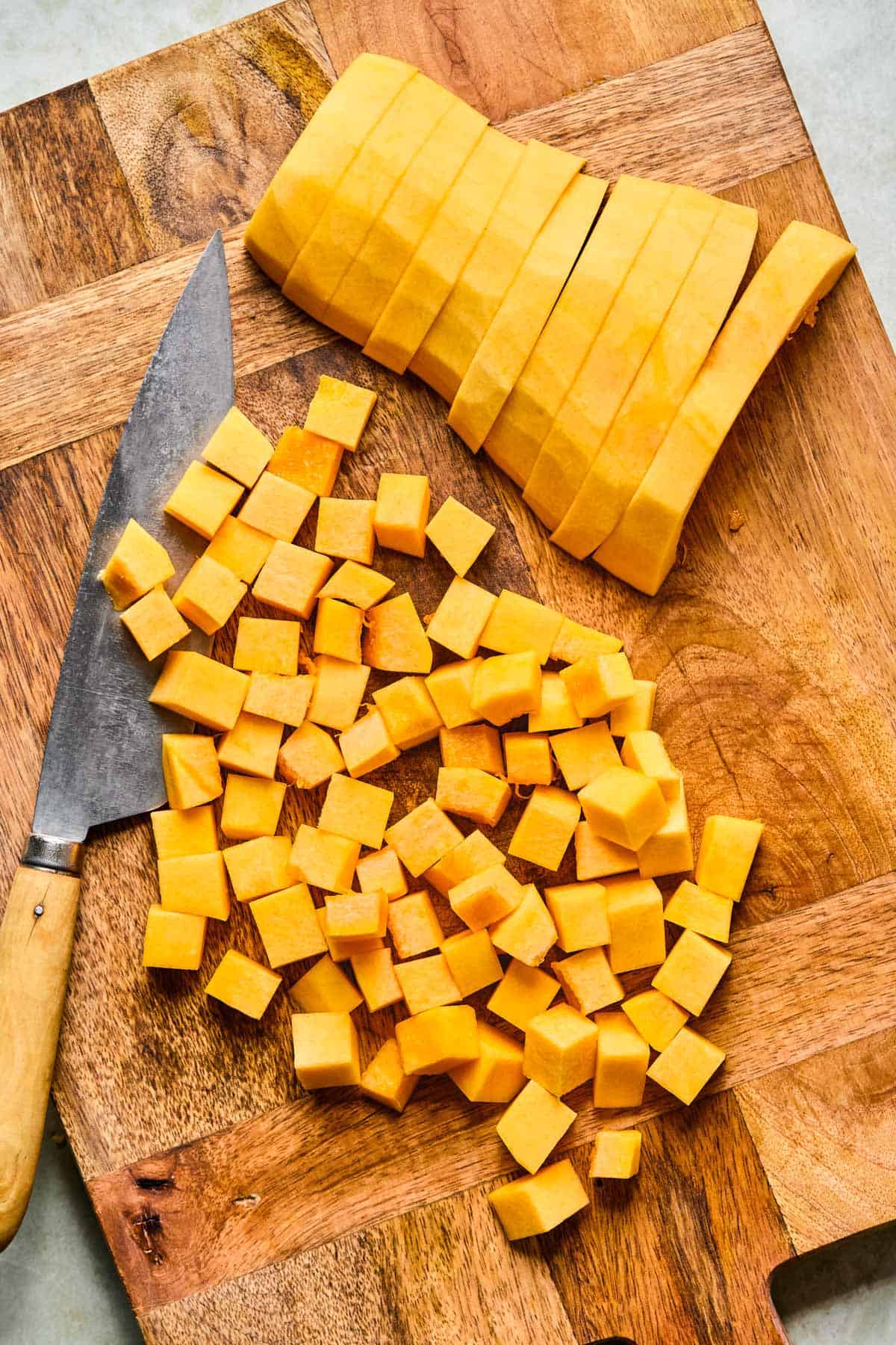 Peeled butternut squash sliced and cubed on a wooden cutting board with a chef’s knife.