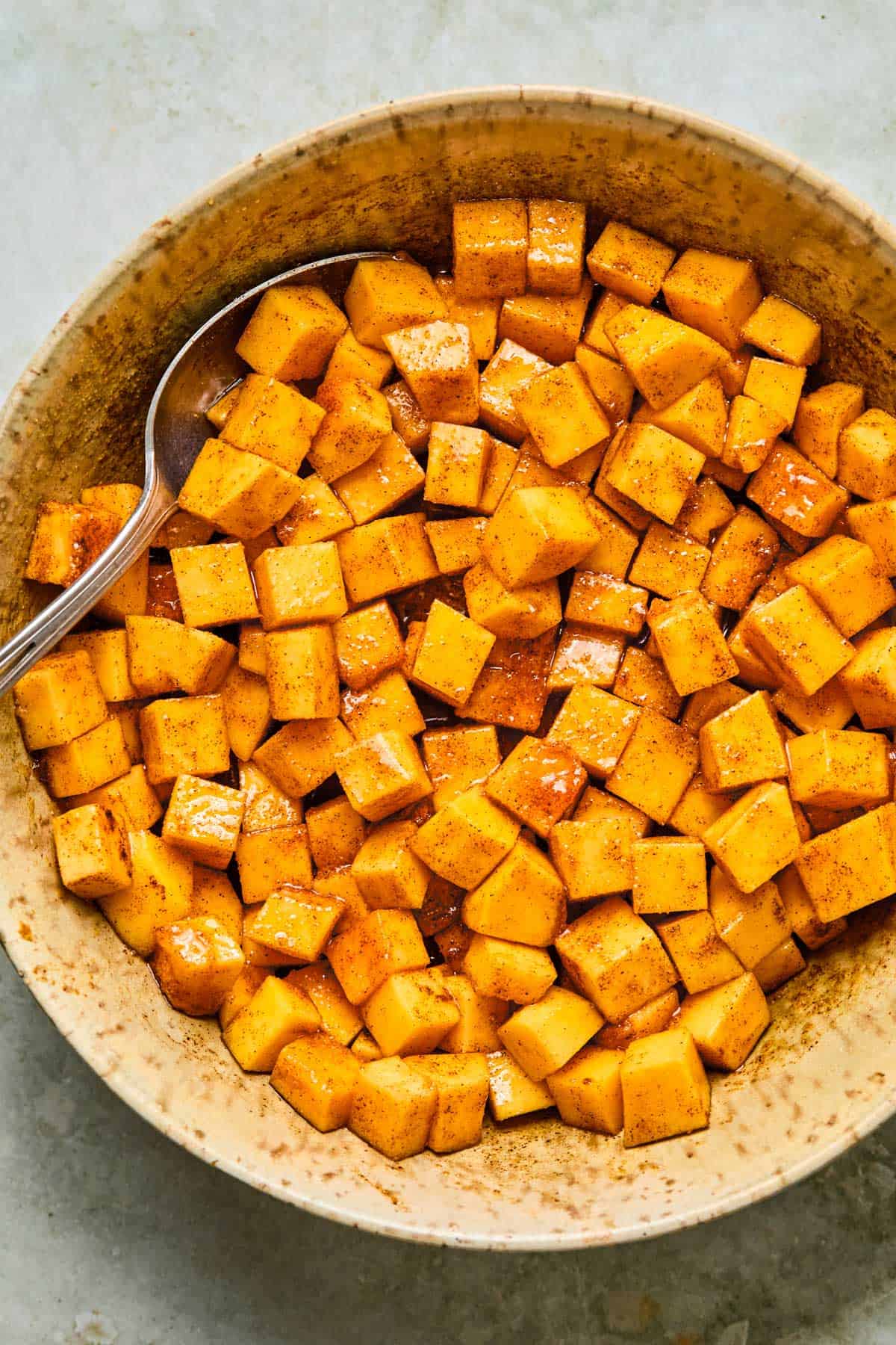 Cubed butternut squash tossed with olive oil, maple syrup, and spices in a ceramic mixing bowl with a spoon.
