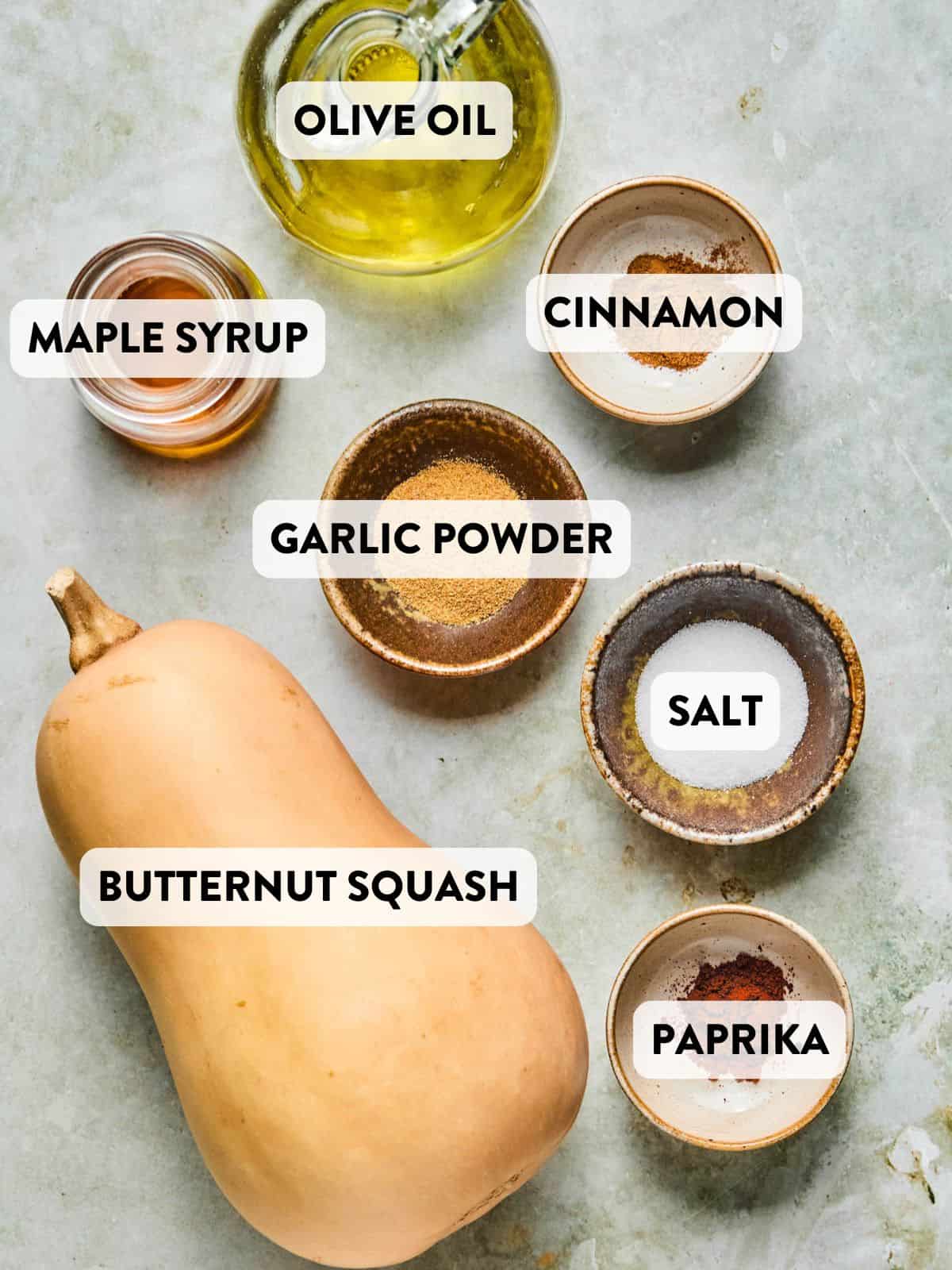 Raw butternut squash, olive oil, maple syrup, garlic powder, paprika, cinnamon, and salt arranged on a marble surface in small bowls.