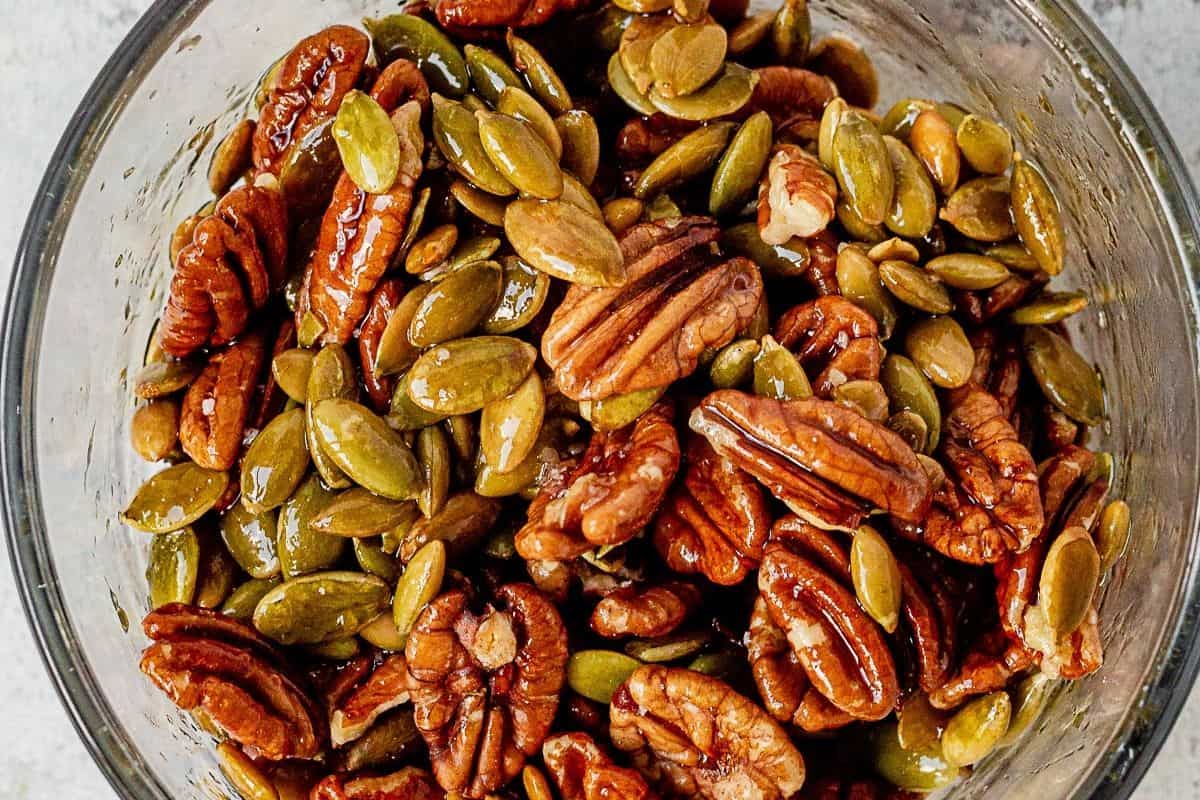 maple and olive oil coating pecans and pumpkin seeds in a bowl