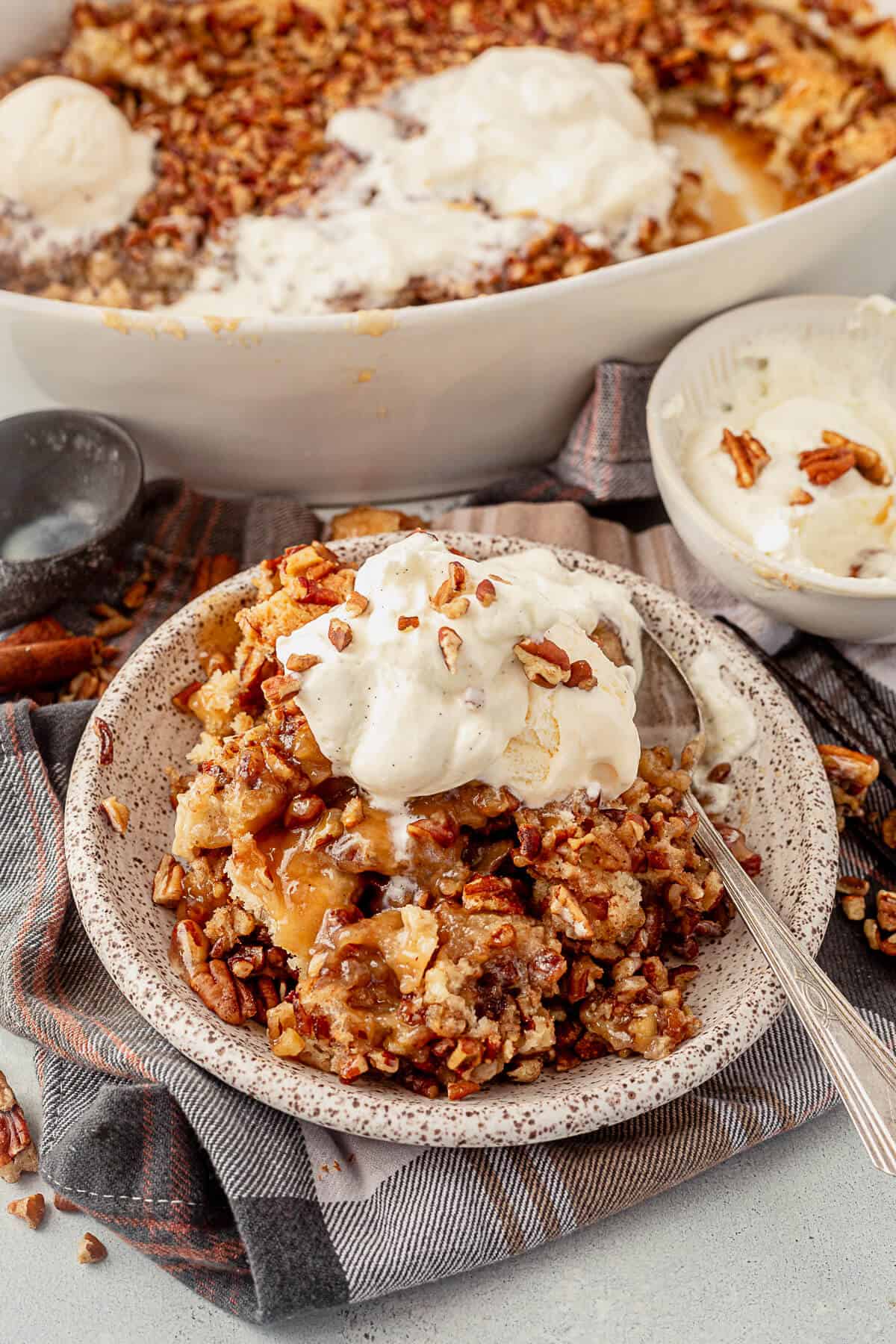 a plate full of pecan pie cobbler with whipped cream and a spoon next to the large dish of cobbler