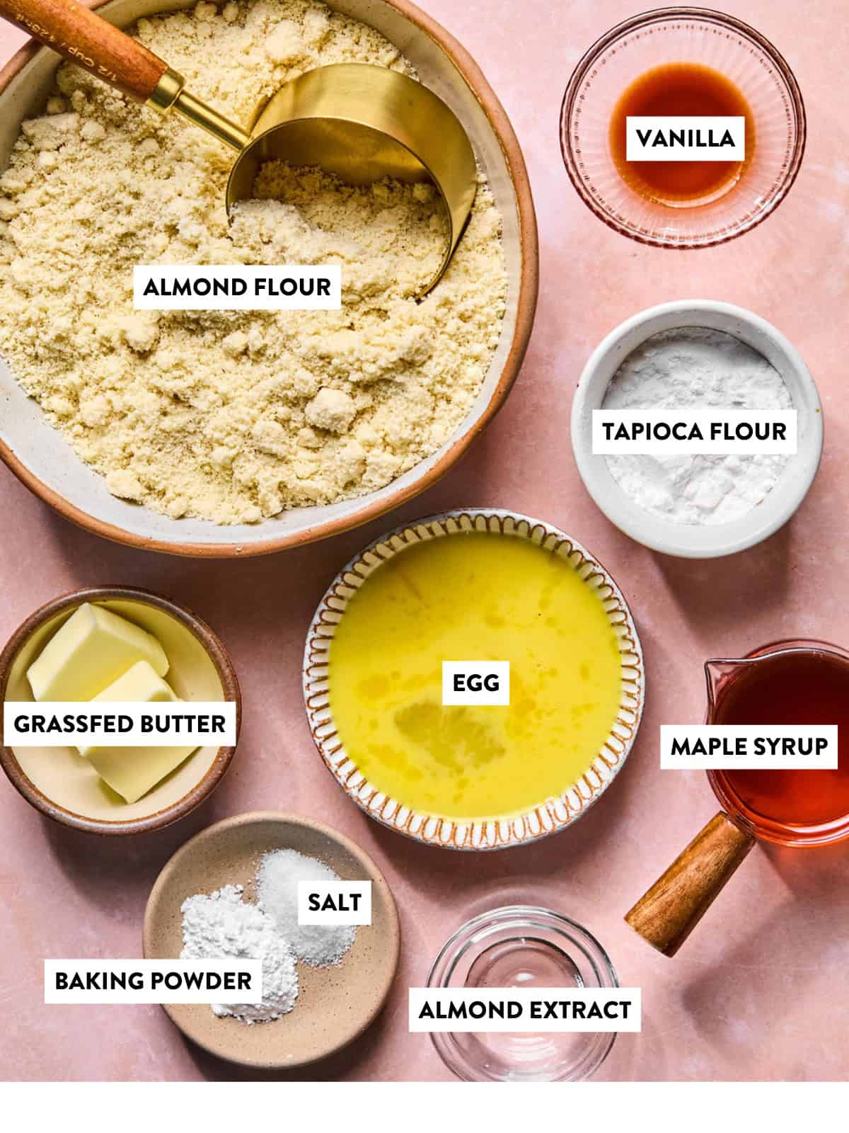 Top-down view of labeled baking ingredients for gluten free biscotti: almond flour, tapioca flour, vanilla, maple syrup, almond extract, salt, baking powder, melted grassfed butter, and a beaten egg, all arranged on a pink surface.