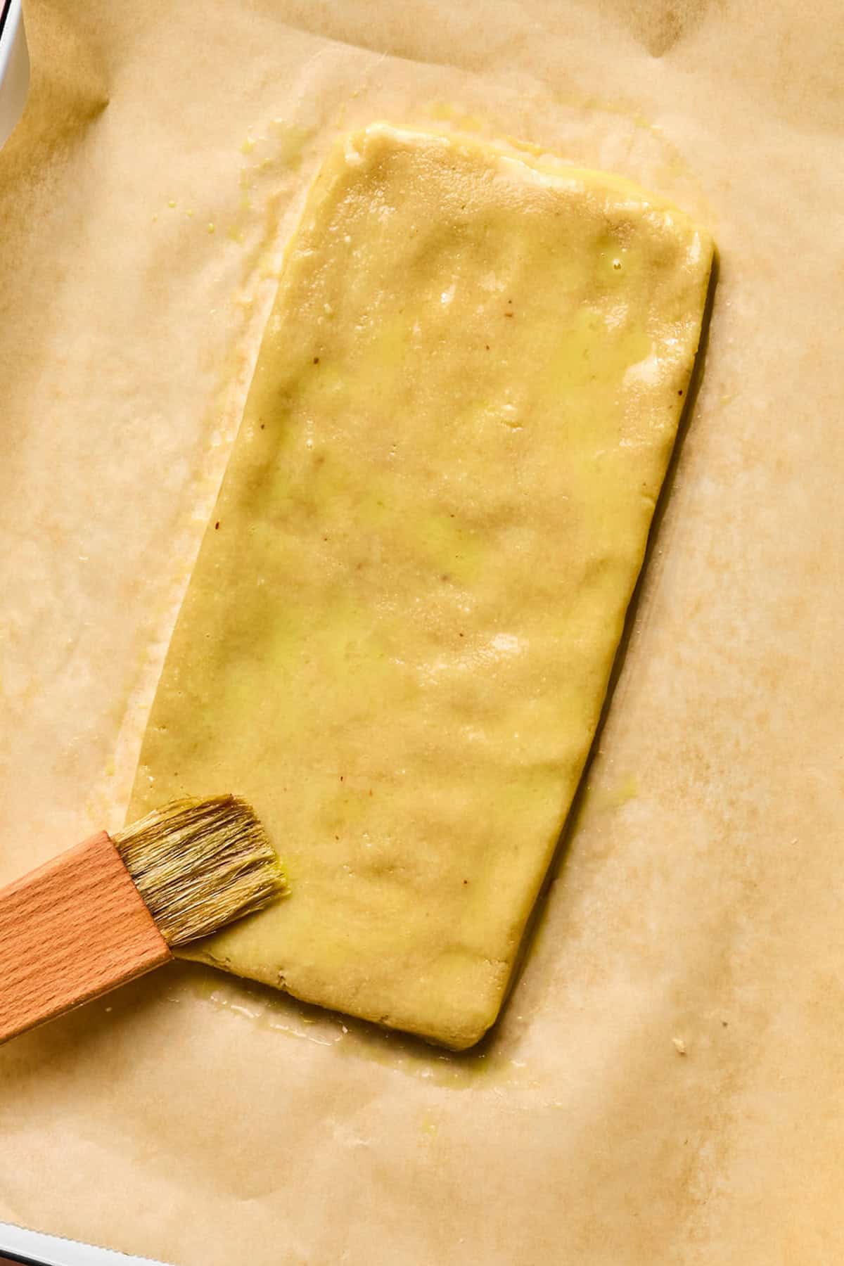 A pastry rectangle on parchment paper being brushed with egg wash using a wooden pastry brush, similar to the careful preparation of gluten free biscotti.