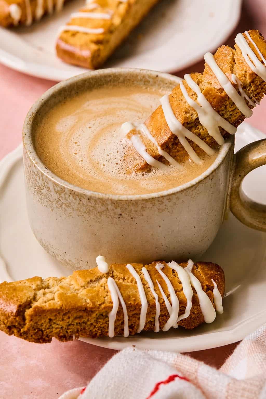 gluten free almond flour biscotti resting in a hot latte on a plate with another biscotti piece resting on the plate.