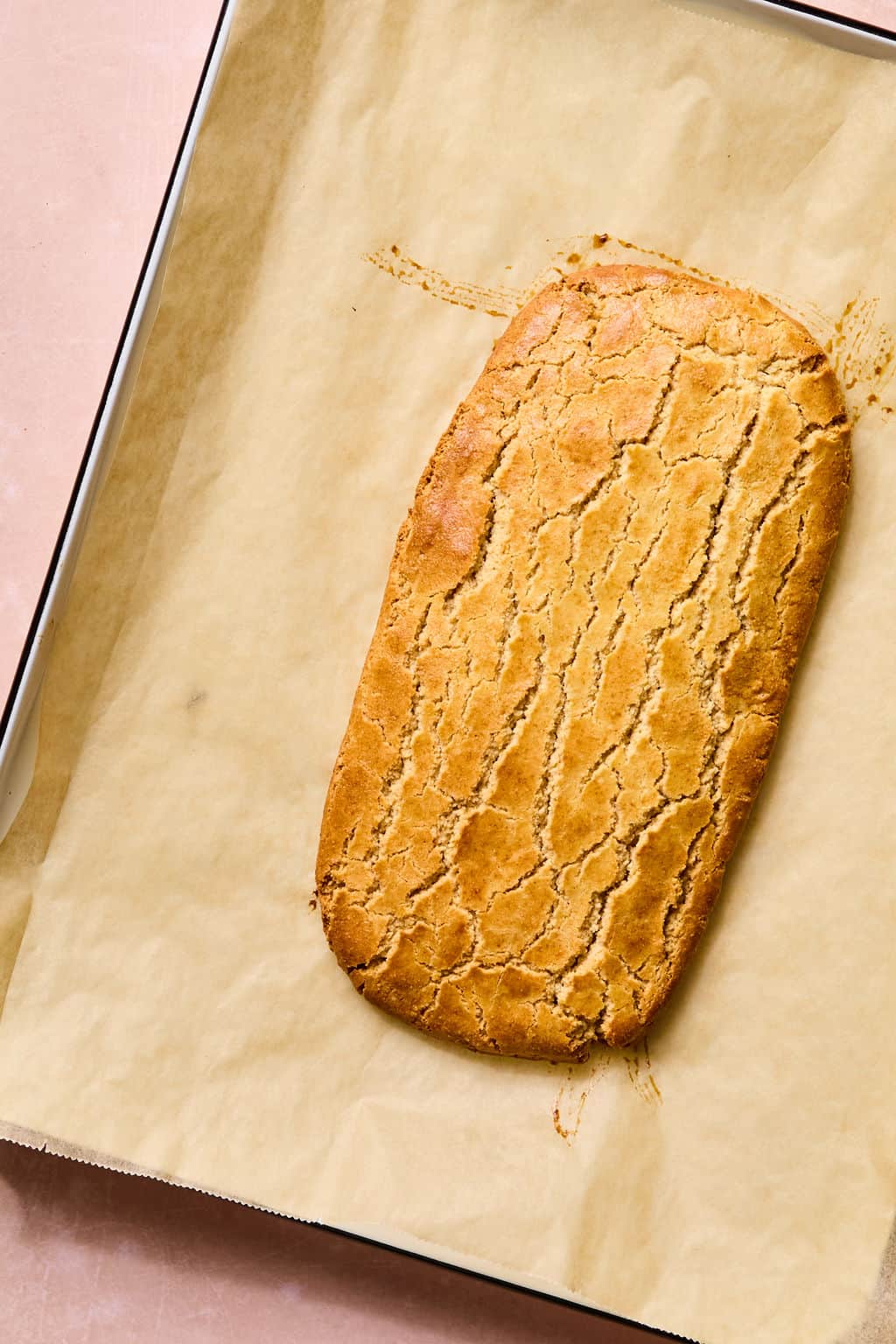 A rectangular loaf of golden brown almond flour biscotti with a golden surface sits on parchment paper in a baking tray.