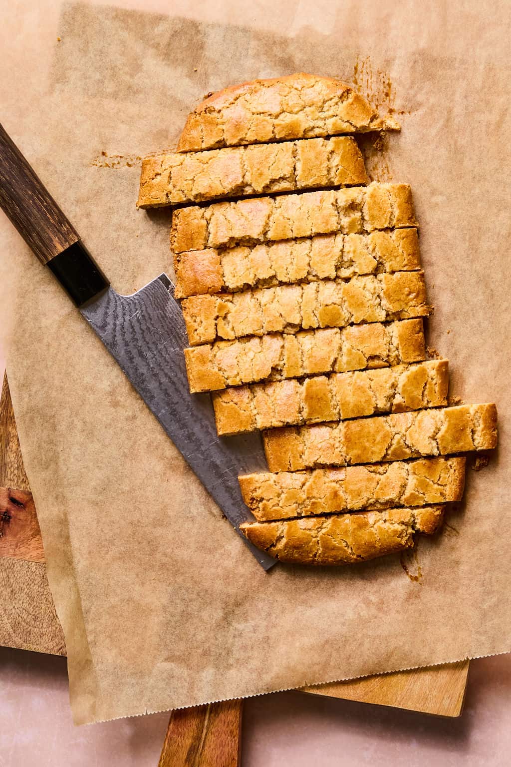 Several slices of golden brown gluten free almond flour biscotti are arranged on parchment paper next to a knife with a dark handle, all placed on a wooden cutting board.