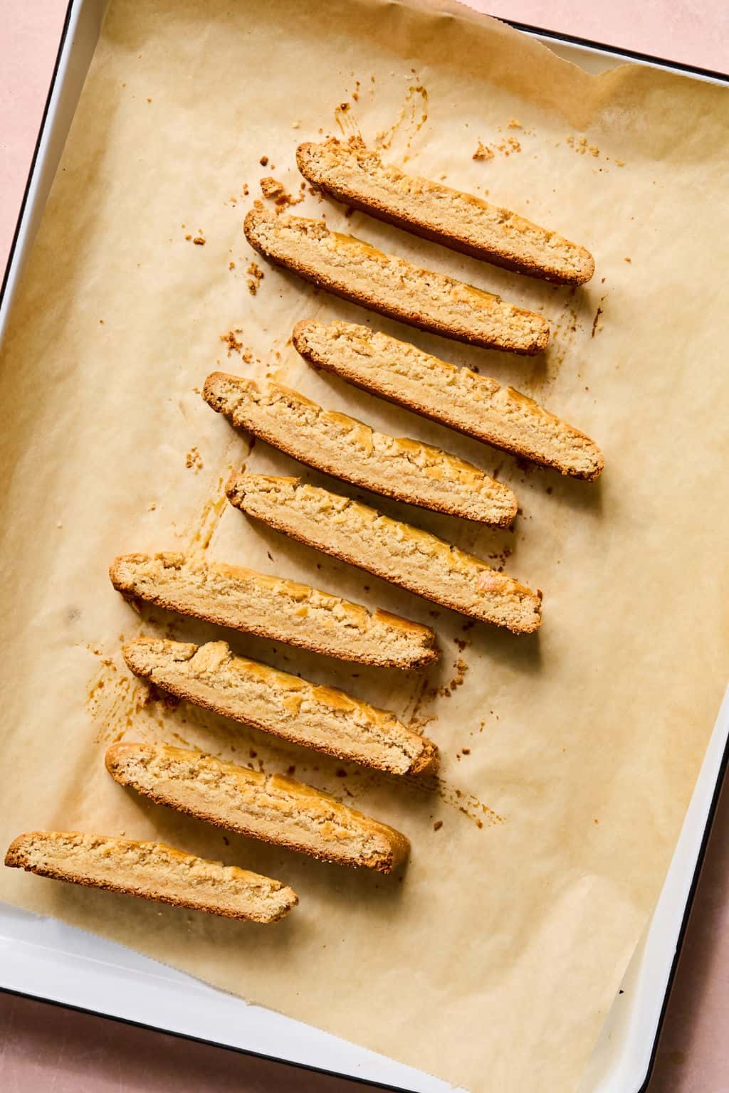 Eight gluten free biscotti pieces are arranged in a diagonal line on a parchment-lined baking sheet, viewed from above. The biscotti are golden brown and crumbly, with small crumbs scattered around them.