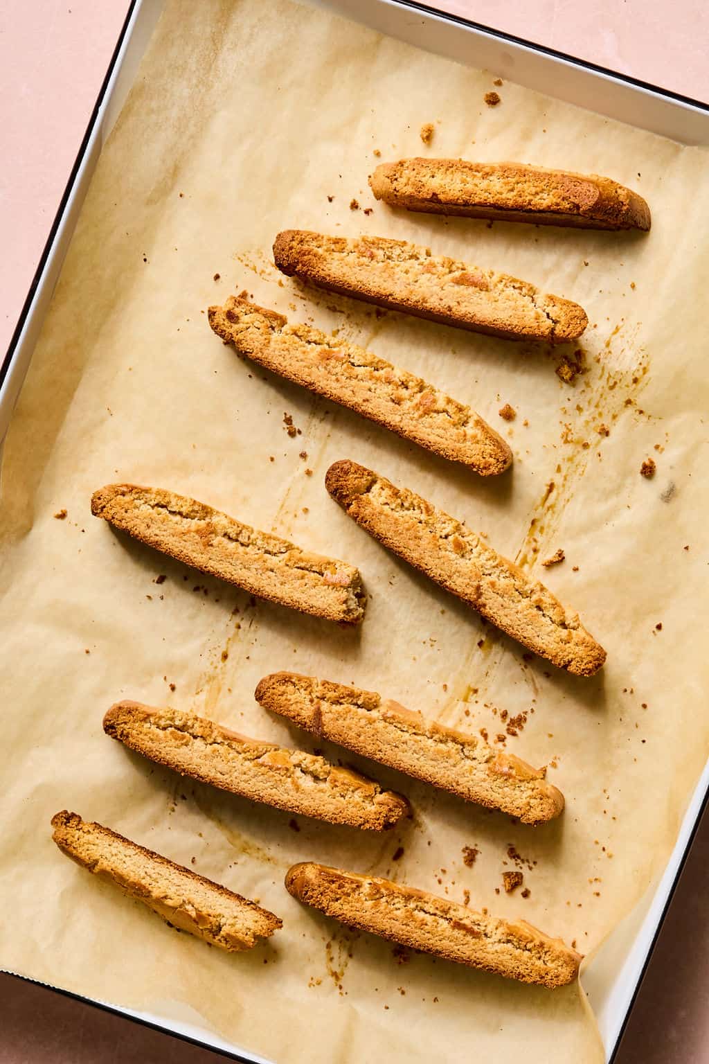 Seven golden brown gluten free biscotti cookies are arranged on a baking tray lined with parchment paper. The tray rests on a light pink surface, and a few cookie crumbs are scattered around.