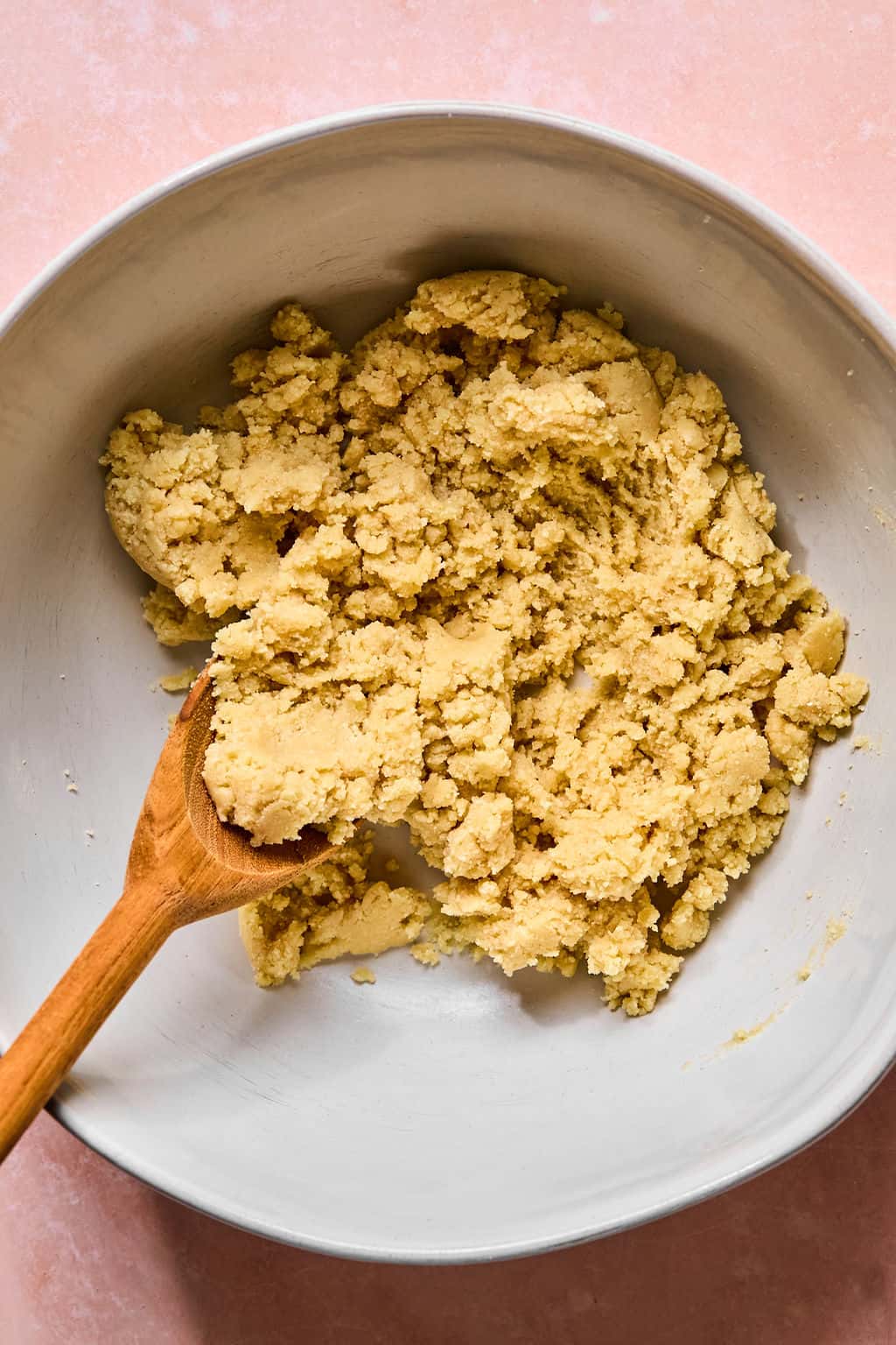A mixing bowl containing crumbly gluten free biscotti cookie dough, being stirred with a wooden spoon, sits on a light pink surface.
