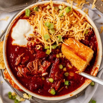 crock pot steak chili in a bowl with cheese, cornbread, sour cream, and green onions