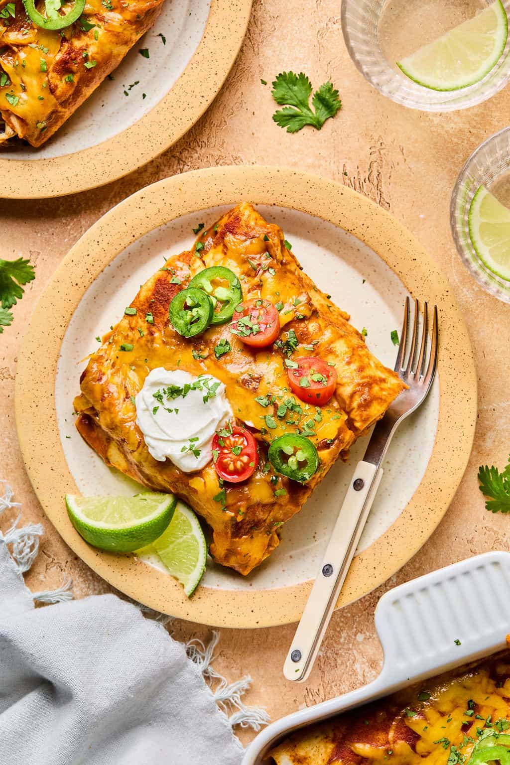 A plate with healthy enchiladas topped with sliced jalapeños, cherry tomatoes, and sour cream, served with lime wedges and a fork, sits on a beige table next to a refreshing glass of water with lime.