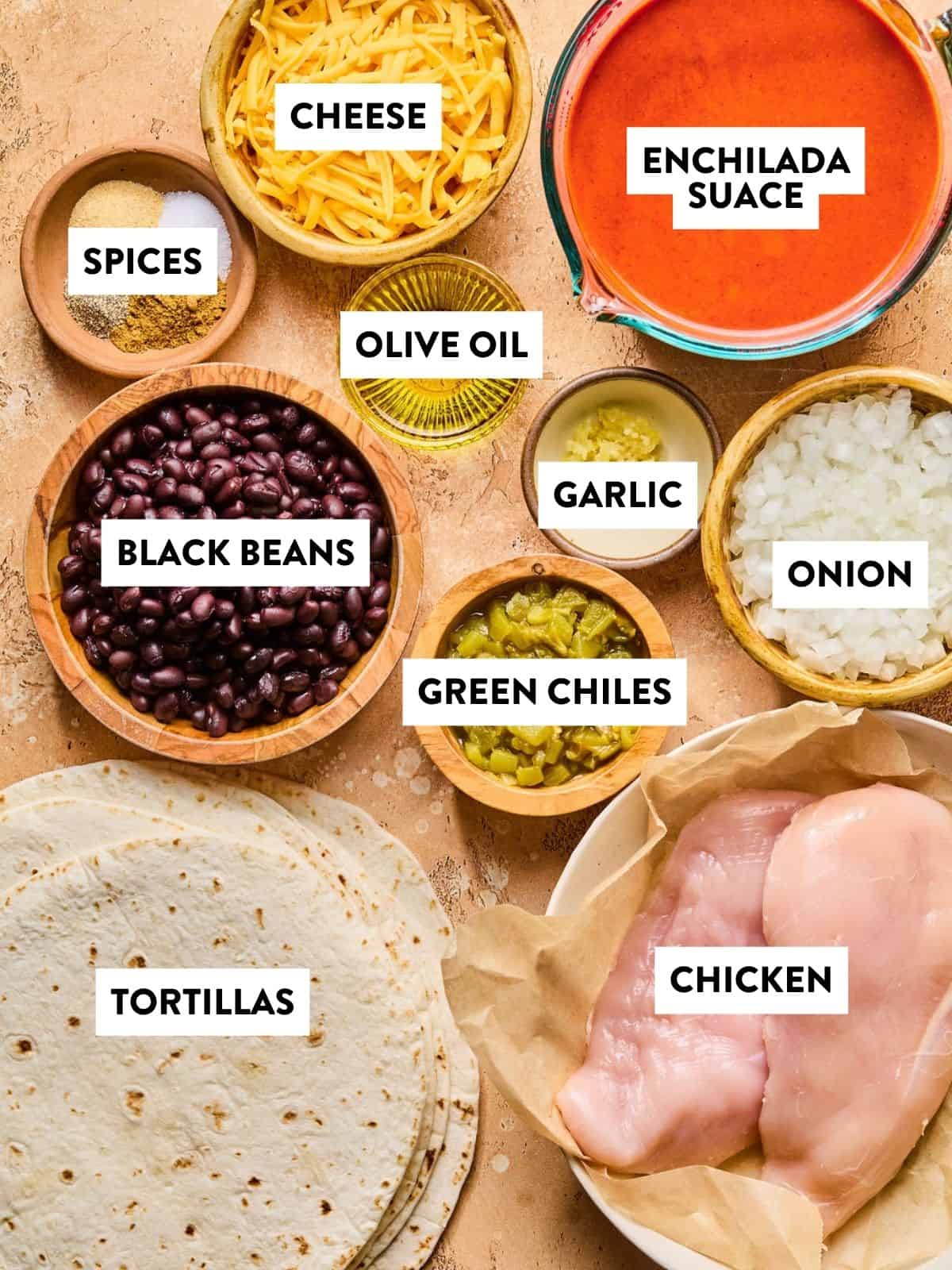 Ingredients for healthy enchiladas are arranged on a counter, including labeled bowls of cheese, enchilada sauce, spices, olive oil, black beans, garlic, onion, green chiles, tortillas, and raw chicken breast.