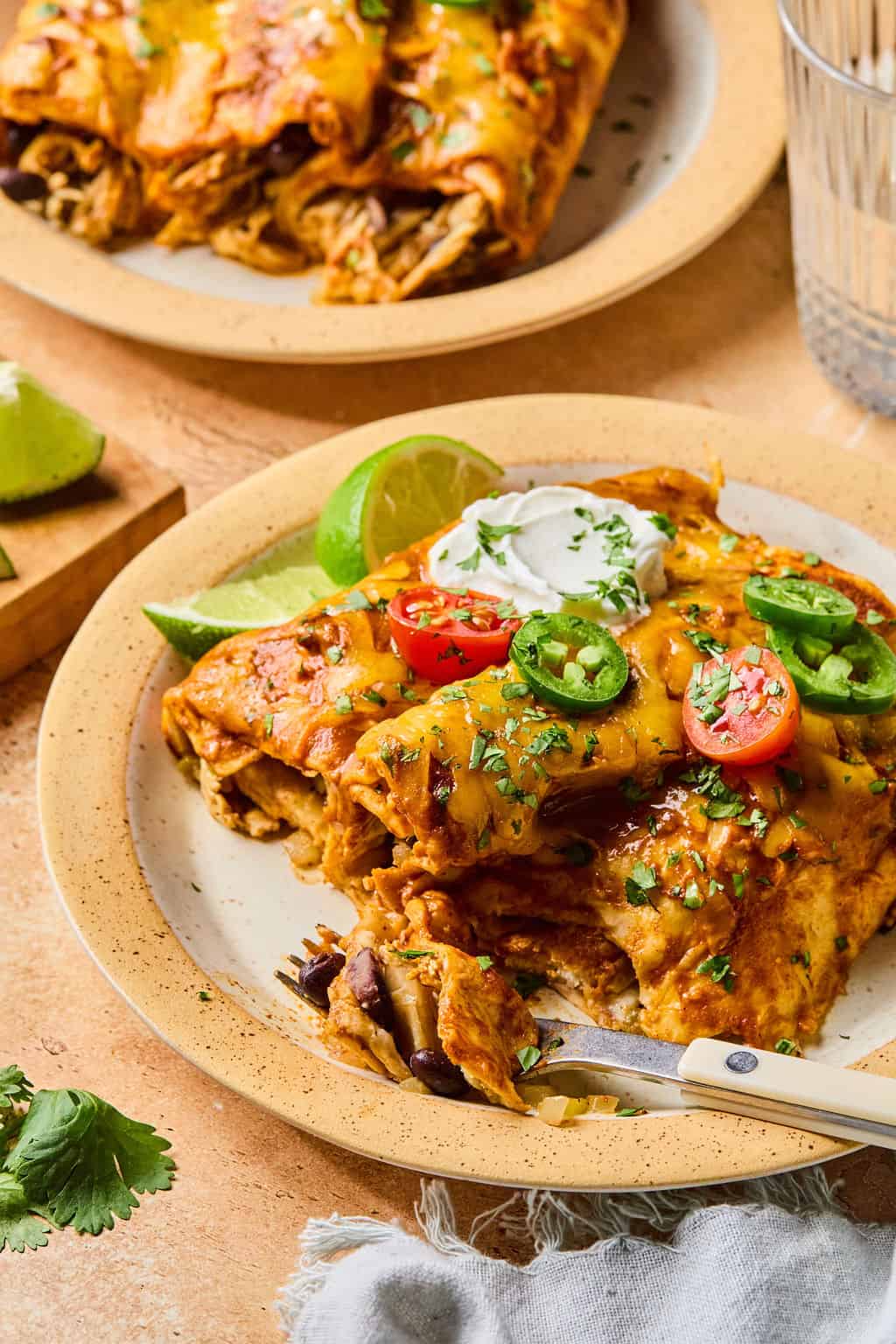 A plate of cheesy, healthy enchiladas topped with sour cream, sliced jalapeños, cherry tomatoes, lime wedges, and chopped cilantro, with a fork taking a bite. Another plate of enchiladas and a glass of water are in the background.