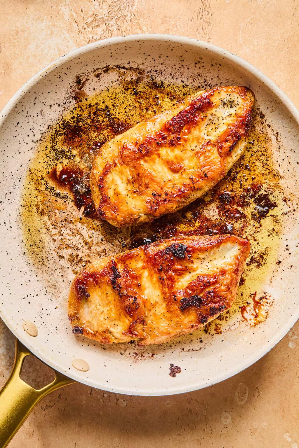 Two golden-brown, seared chicken breasts cooking in a lightly oiled skillet, perfect for adding to healthy enchiladas, with seasoning and crispy edges visible on a beige surface.