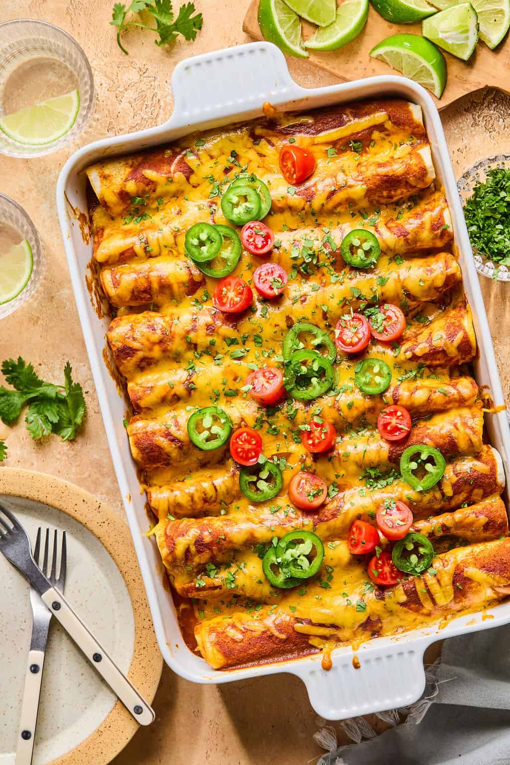 A baking dish filled with healthy enchiladas topped with sliced jalapeños, cherry tomatoes, and chopped herbs. Plates, cutlery, lime wedges, and fresh cilantro are arranged nearby on a tan surface.