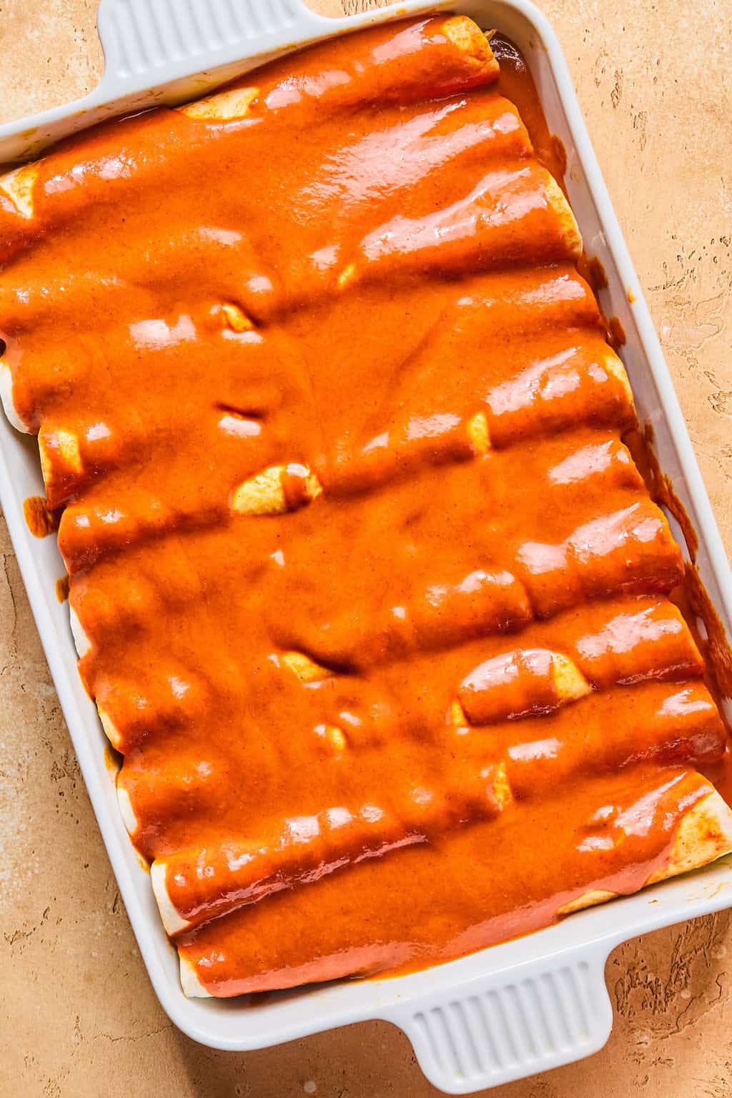 A white rectangular baking dish filled with healthy enchiladas, each rolled and topped with a generous layer of red enchilada sauce, ready to be baked.