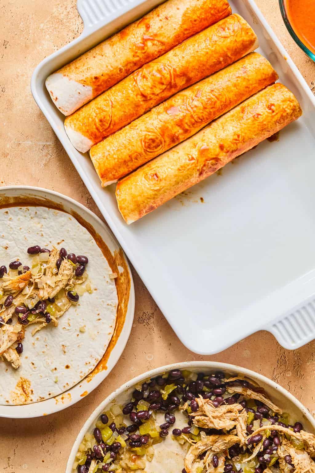Four healthy enchiladas in a white baking dish sit next to bowls with tortillas, shredded chicken, black beans, and green chiles on a tan countertop.