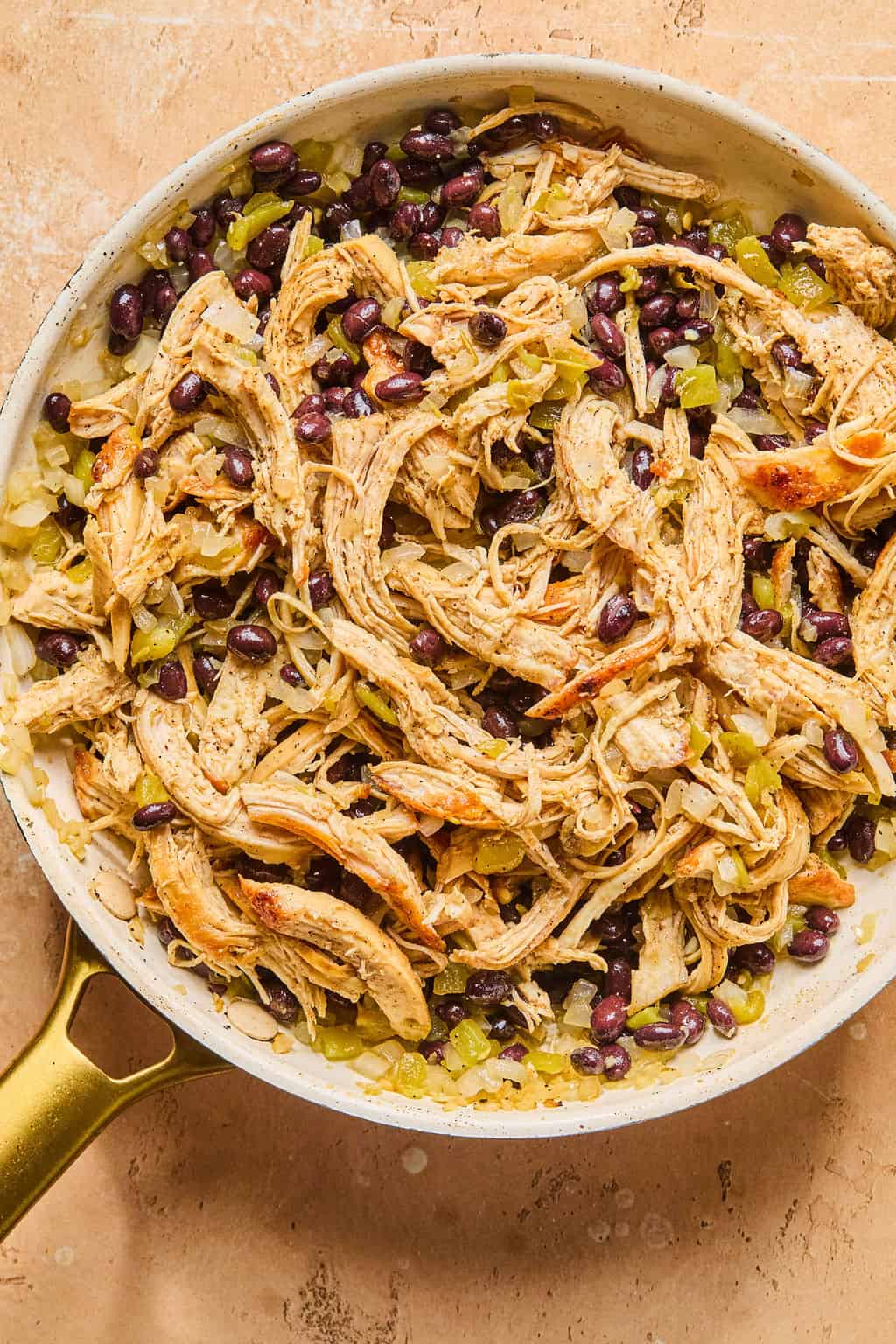 A skillet filled with shredded chicken, black beans, chopped green chilies, and cooked onions, all mixed together on a tan countertop—perfect for making healthy enchiladas.