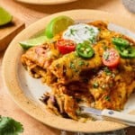A plate of healthy enchiladas topped with sliced cherry tomatoes, jalapeños, cilantro, and a dollop of sour cream, with lime wedges on the side and a fork partially inserted into the food.