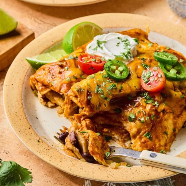 A plate of healthy enchiladas topped with sliced cherry tomatoes, jalapeños, cilantro, and a dollop of sour cream, with lime wedges on the side and a fork partially inserted into the food.