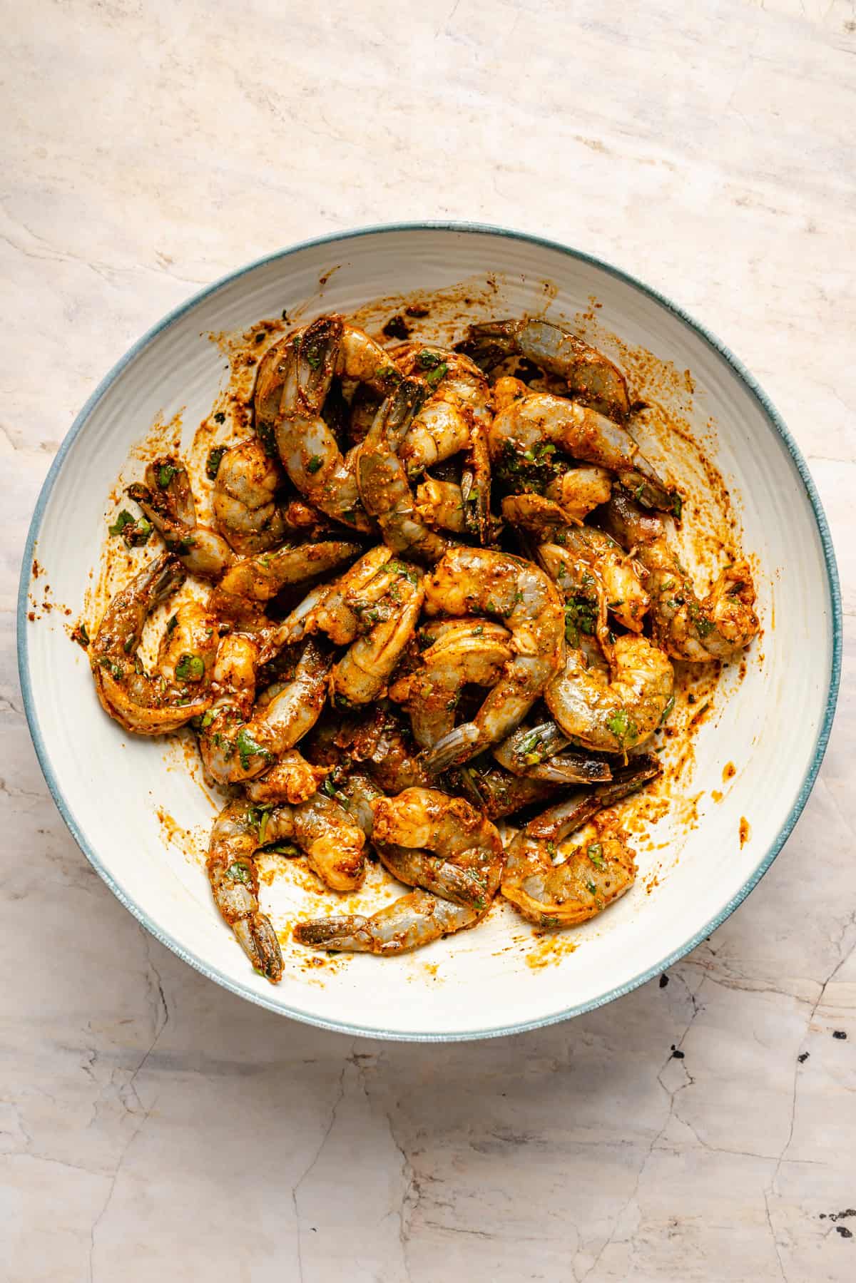 Raw shrimp tossed with spices and herbs in a white bowl, sitting on a light marble surface. The shrimp are coated in an orange marinade and are ready to be cooked—perfect for your next shrimp bowl recipe.