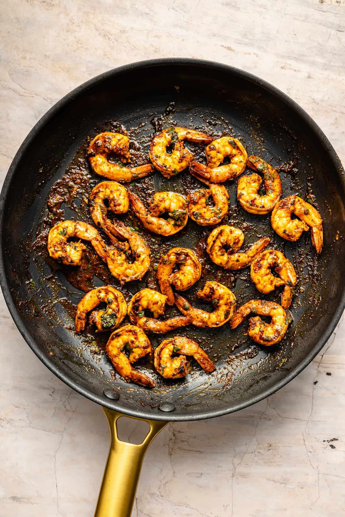 Cooked, seasoned shrimp arranged in a black skillet with a gold handle, sitting on a light marble surface—an irresistible shrimp bowl recipe inspiration. The shrimp are slightly charred and coated in spices, creating a rustic and appetizing appearance.