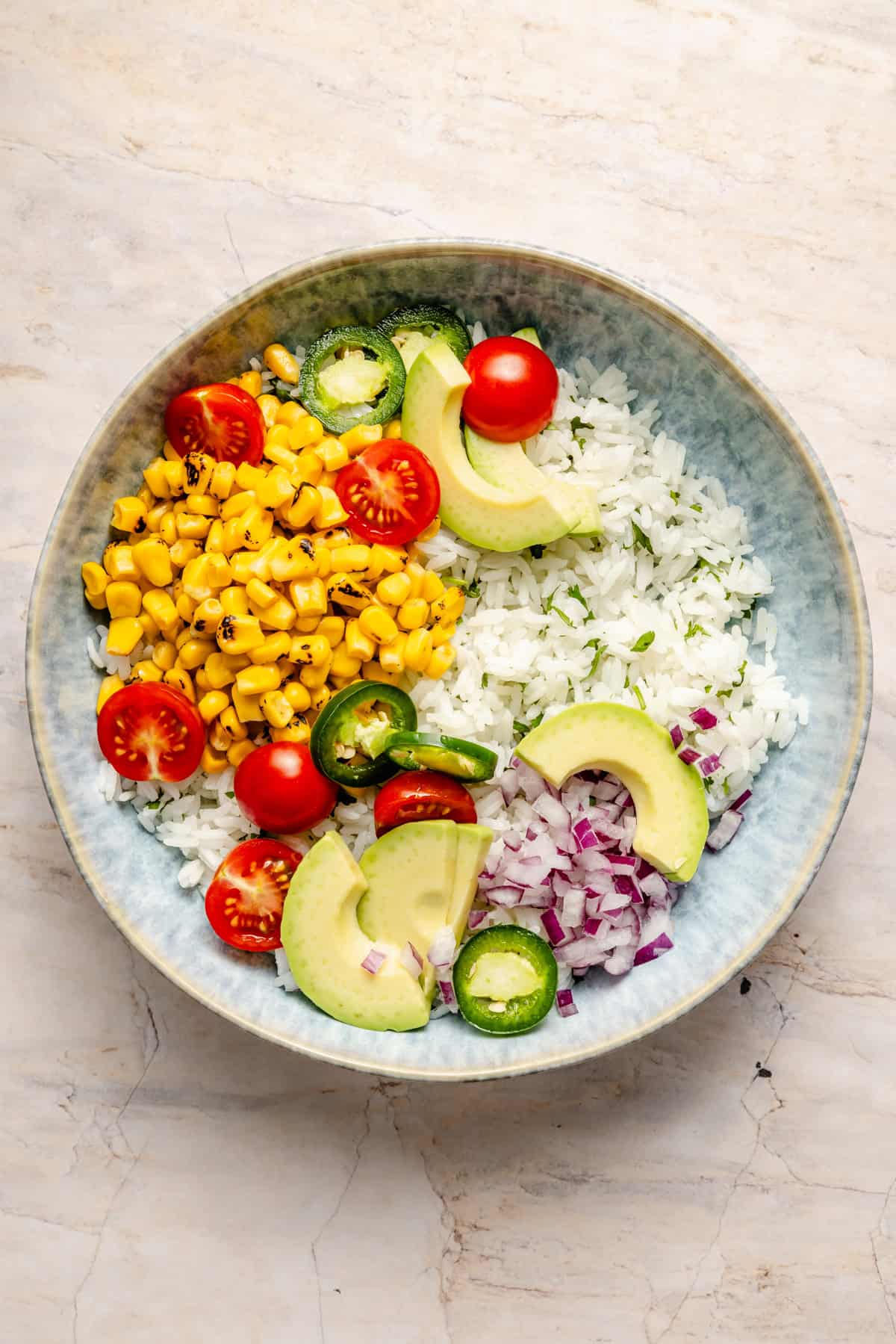 A bowl with chopped grilled corn, halved cherry tomatoes, sliced avocado, jalapeño, red onion, and white onion on a marble surface—perfect as a vibrant base for your favorite shrimp bowl recipe.