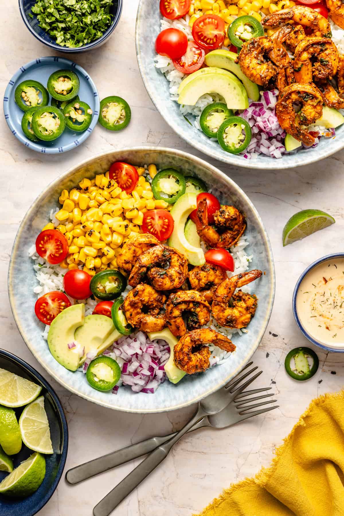 Two shrimp bowls with rice, grilled shrimp, corn, cherry tomatoes, avocado, red onion, and jalapeño slices showcase a vibrant shrimp bowl recipe. Small bowls with lime wedges, sliced jalapeños, chopped herbs, and creamy sauce are nearby on a white surface.
