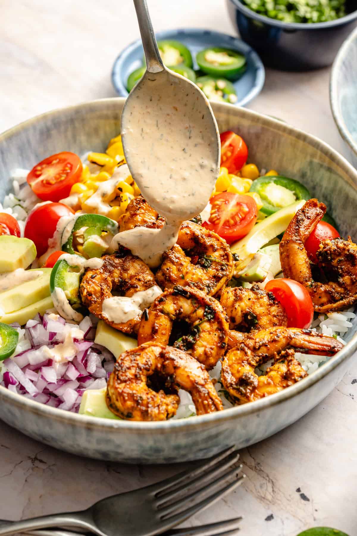 A vibrant shrimp bowl recipe featuring a bowl of rice topped with grilled shrimp, avocado, cherry tomatoes, red onion, corn, and jalapeño slices, with creamy dressing being drizzled on top. A fork and knife are placed in front of the bowl.