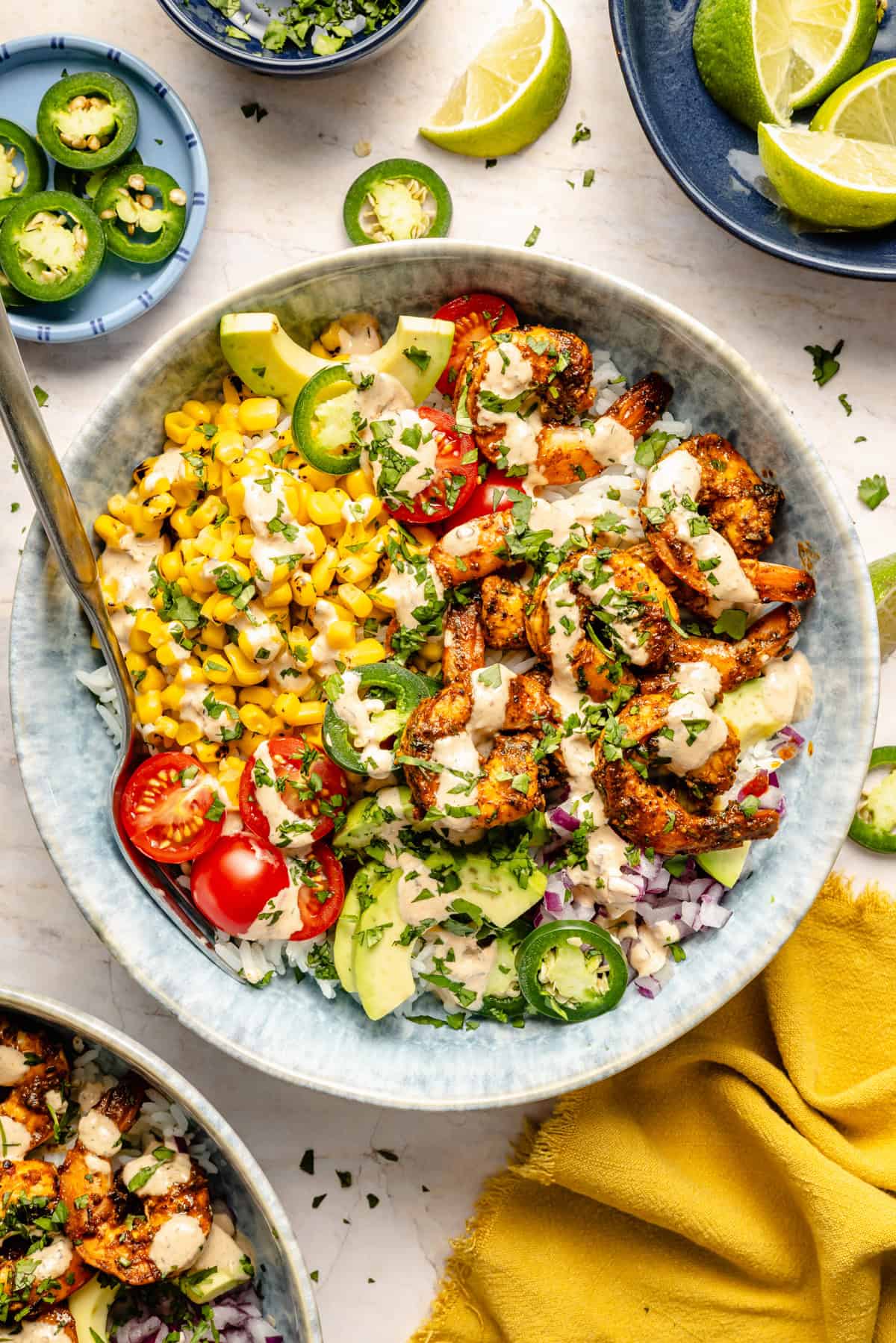 A delicious shrimp bowl recipe featuring grilled shrimp, corn, cherry tomatoes, avocado slices, red onion, jalapeño, and cilantro drizzled with creamy sauce; served with lime wedges and other fresh ingredients nearby.