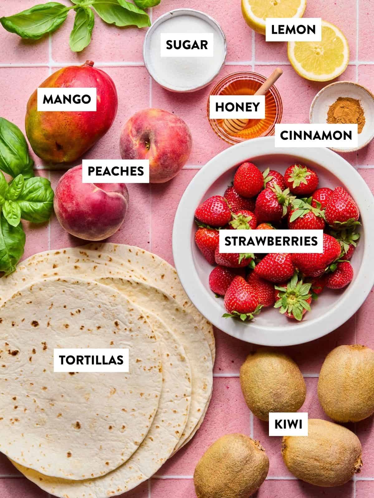Flat lay of ingredients for fruit salsa on a pink tiled surface, including tortillas, strawberries, peaches, mango, kiwi, lemon, basil, plus bowls of sugar, honey, and ground cinnamon—all labeled.