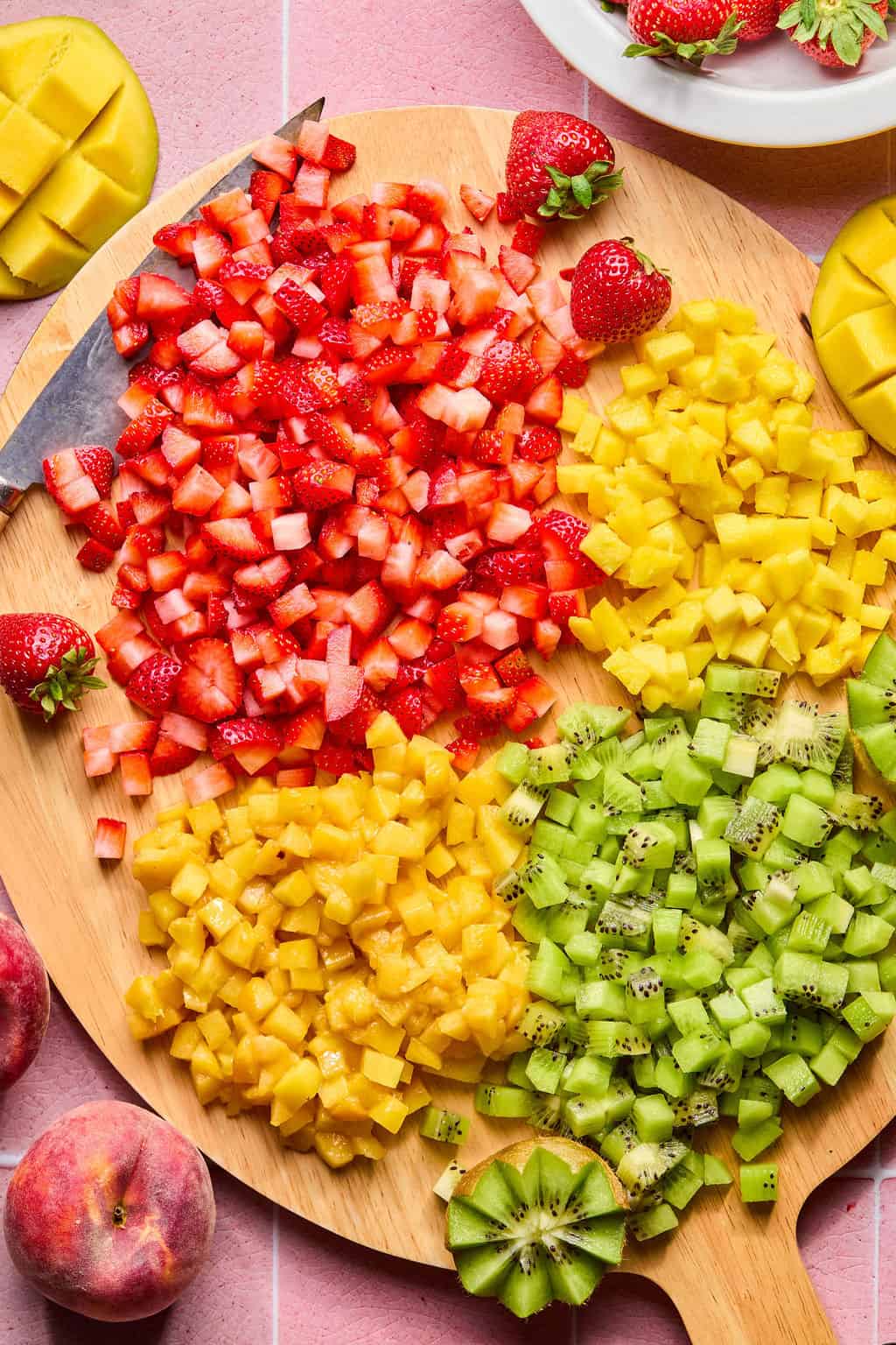 A round wooden board displays neatly diced strawberries, mango, and kiwi in separate sections, perfect for making a fresh fruit salsa. Whole strawberries, mango, kiwi, and peaches are placed around the board on a pink surface.