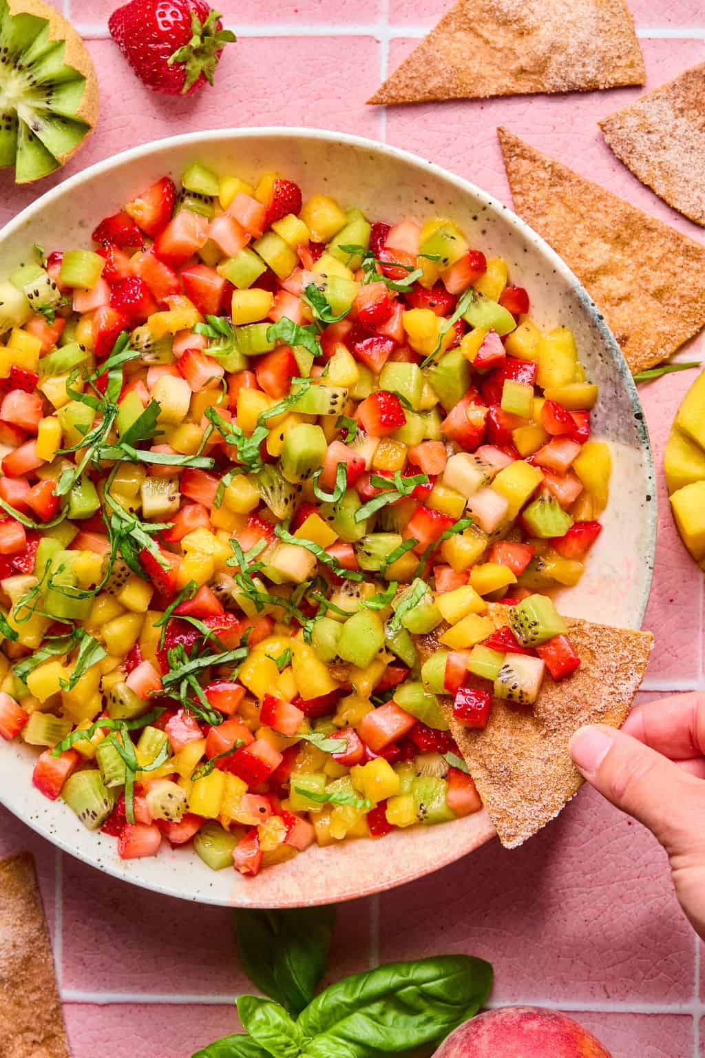 A bowl of colorful fruit salsa made with diced strawberries, kiwi, and mango sits on a pink tiled surface. A hand dips a cinnamon sugar tortilla chip into the vibrant fruit salsa. Fresh fruit and chips are scattered around the bowl.