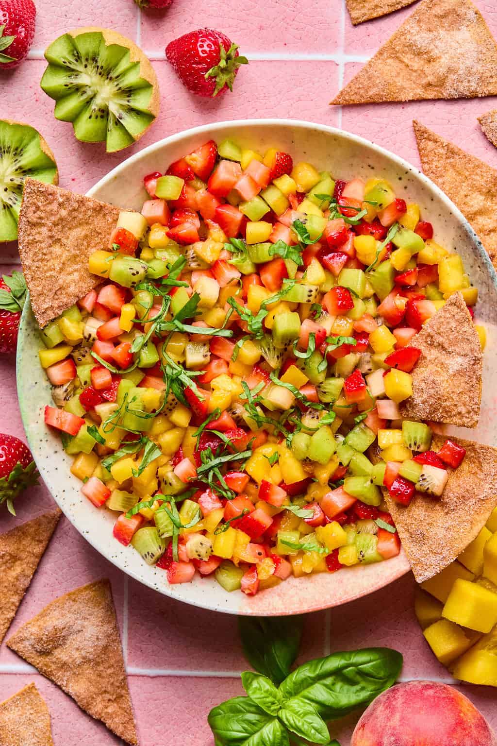 A bowl of vibrant fruit salsa made with diced strawberries, kiwi, mango, and herbs is served with crispy cinnamon tortilla chips on a pink tiled surface. Sliced strawberries, kiwi, basil, and peach are scattered around.