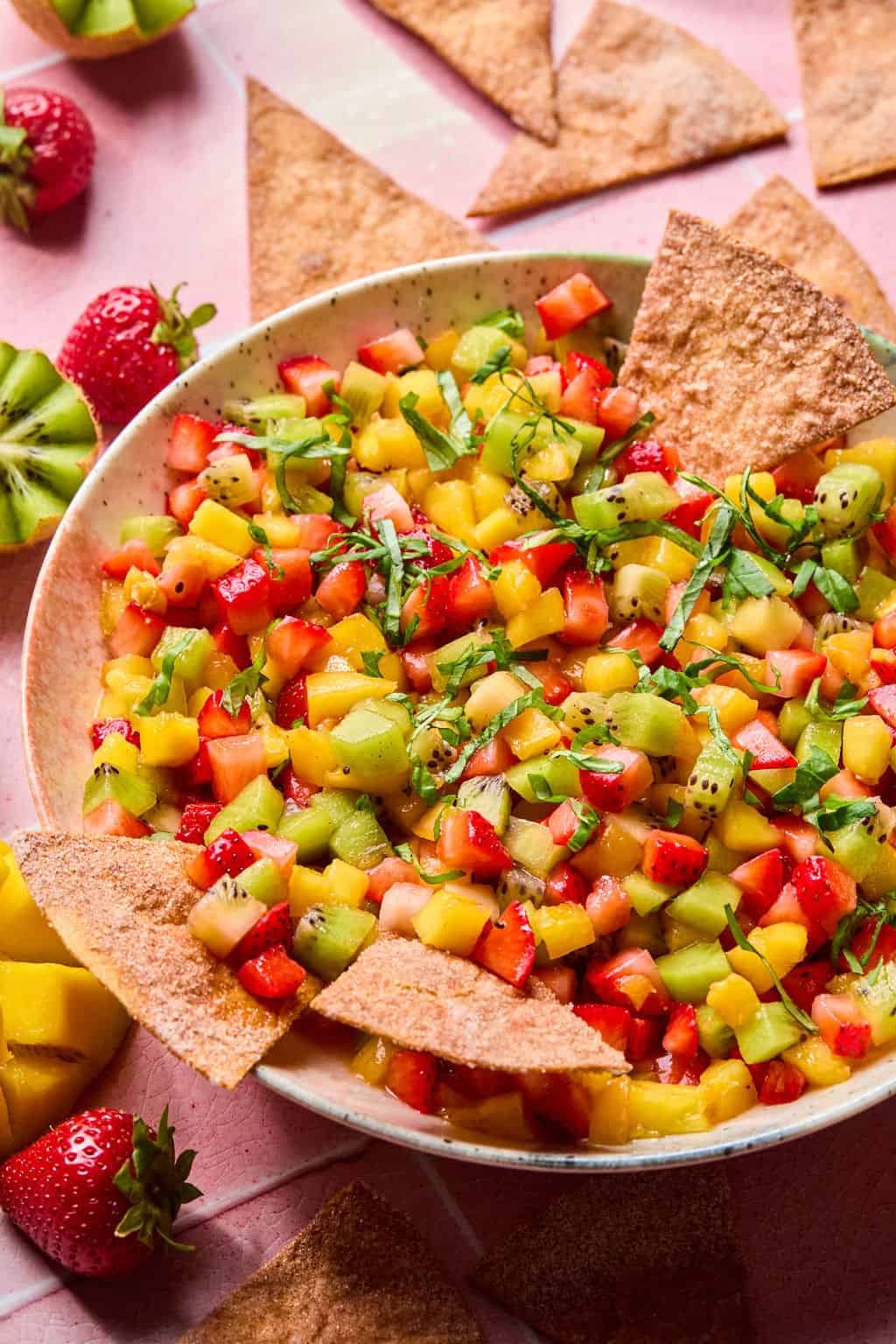 A vibrant bowl of fruit salsa with diced strawberries, kiwi, and mango, garnished with herbs and served with cinnamon-dusted pita chips on a pink surface. Whole strawberries and mango slices complete this fresh fruit salsa spread.