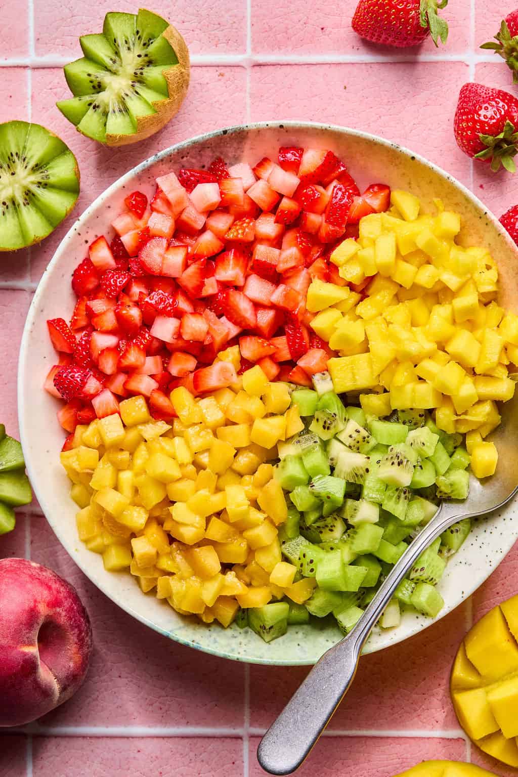 A bowl of fruit salsa made from diced strawberries, mango, and kiwi is arranged in sections with a spoon inside. Whole and halved fruits, including strawberries, kiwi, and a peach, are scattered on a pink tiled surface around the bowl.