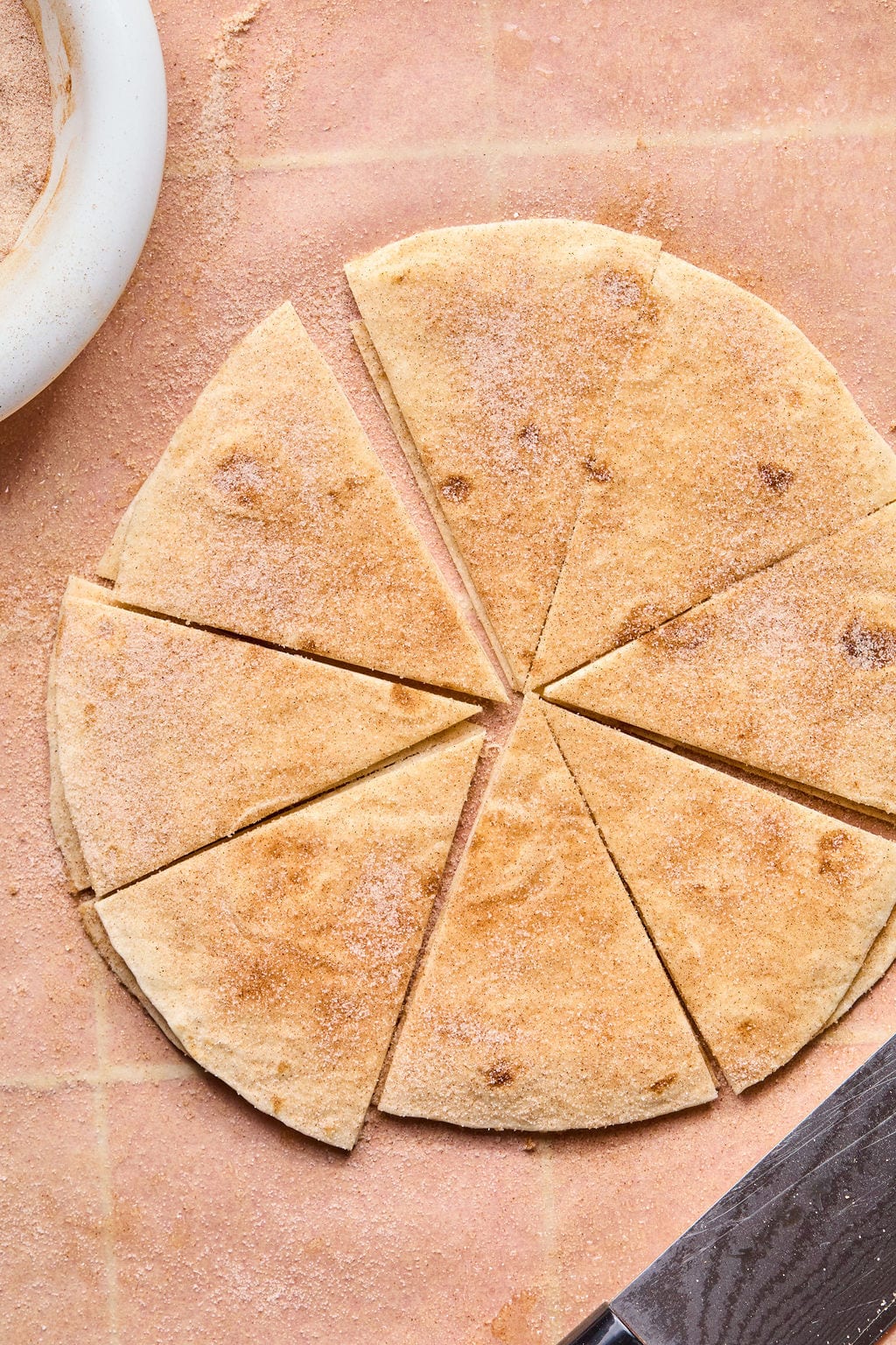 A round tortilla dusted with cinnamon sugar is cut into eight triangular wedges on a pink surface, ready to be paired with fruit salsa, with a knife nearby and a white bowl partially visible in the corner.