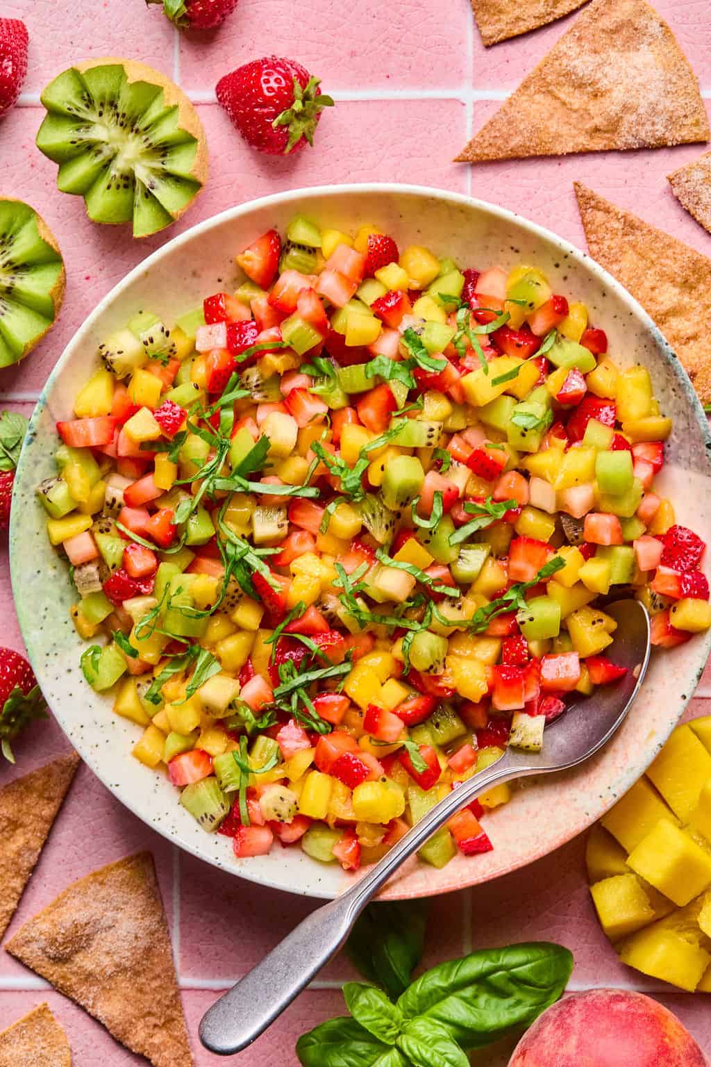 A bowl of vibrant fruit salsa with diced strawberries, kiwi, and mango, garnished with fresh herbs, sits on a pink tiled surface. Surrounded by fruit slices and cinnamon-sugar tortilla chips, a spoon rests in the fruity salsa bowl.