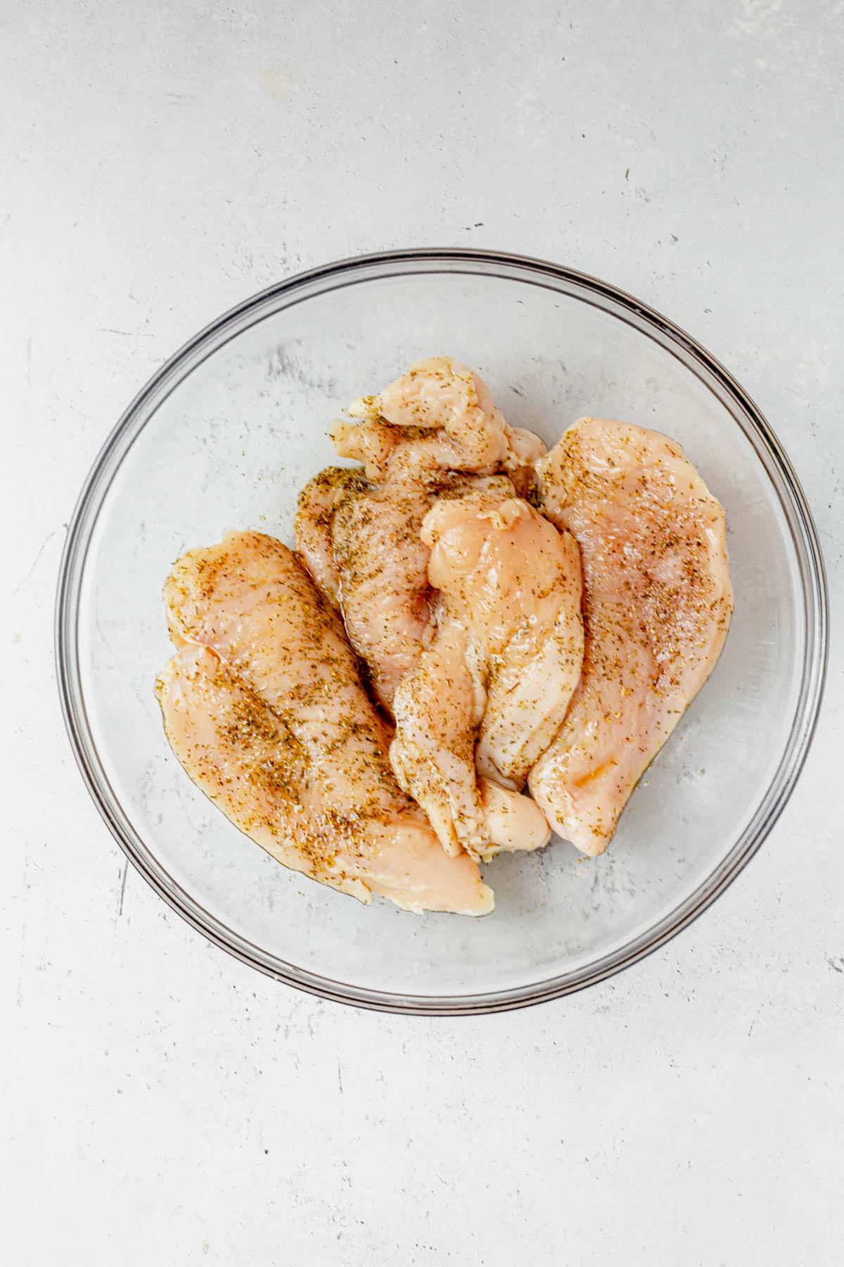 Raw, seasoned chicken breasts in a clear glass bowl on a light gray surface. Coated with visible spices and herbs, these are perfect for making grilled chicken margherita or marinating before cooking.