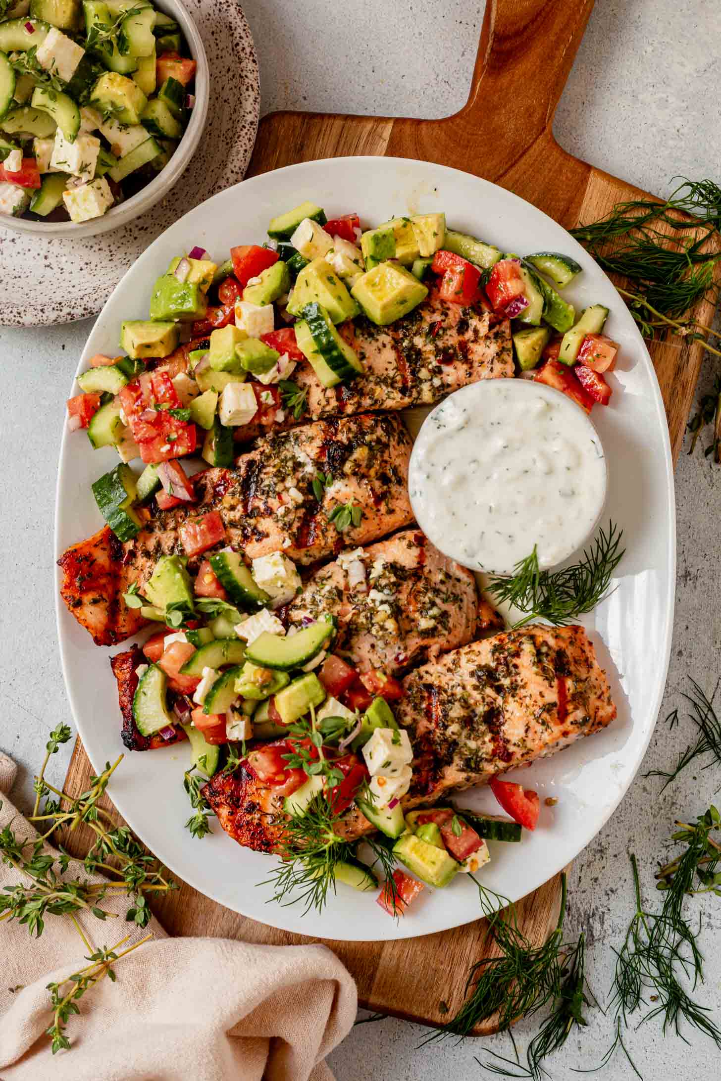 A white plate with Greek salmon filets, herb-grilled and topped with a fresh cucumber, tomato, and avocado salad, served with a side of creamy white dipping sauce and garnished with fresh dill.