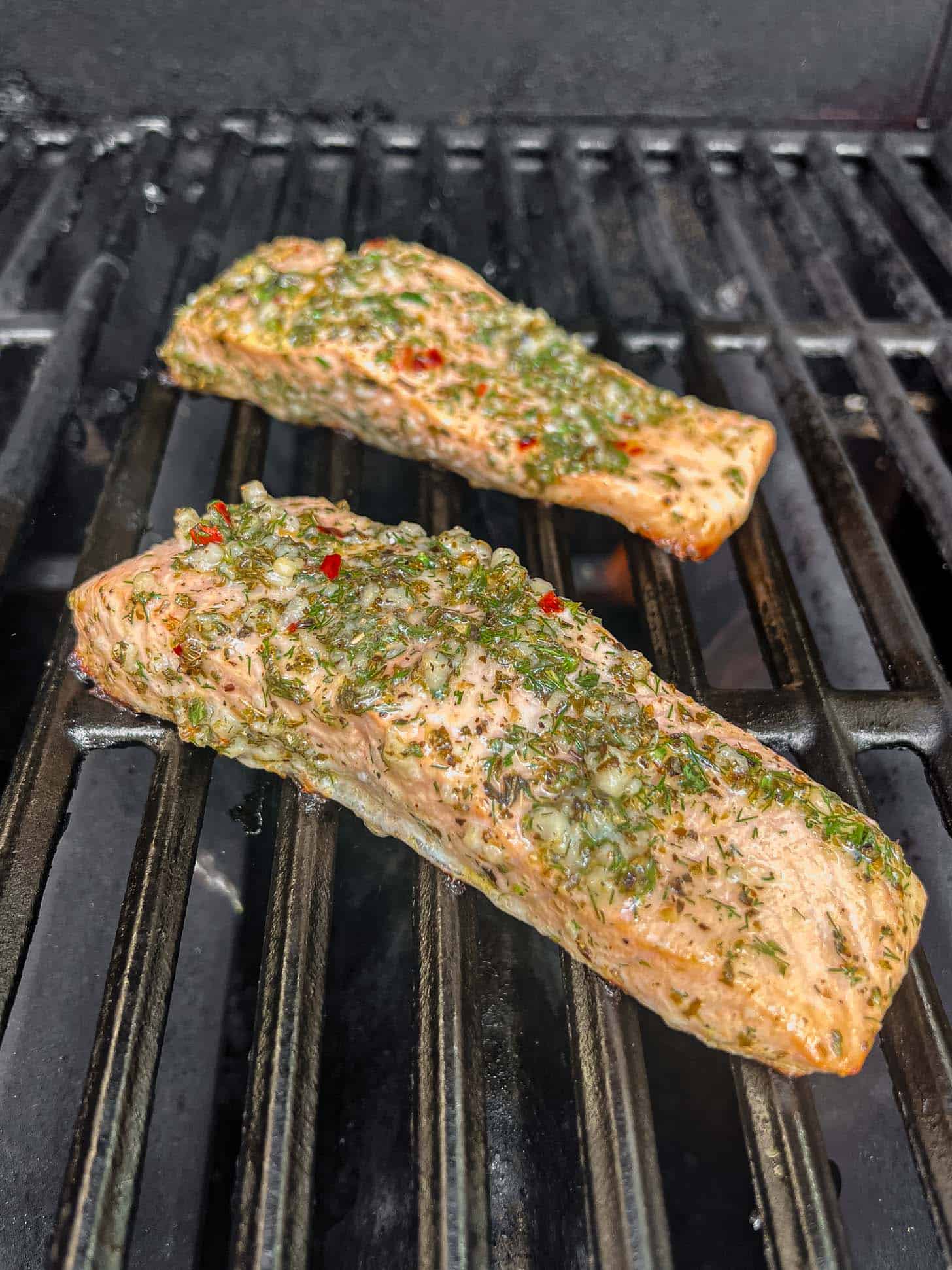 Two seasoned Greek salmon fillets with herbs and spices are grilling on a barbecue, featuring visible grill marks and a smoky background.