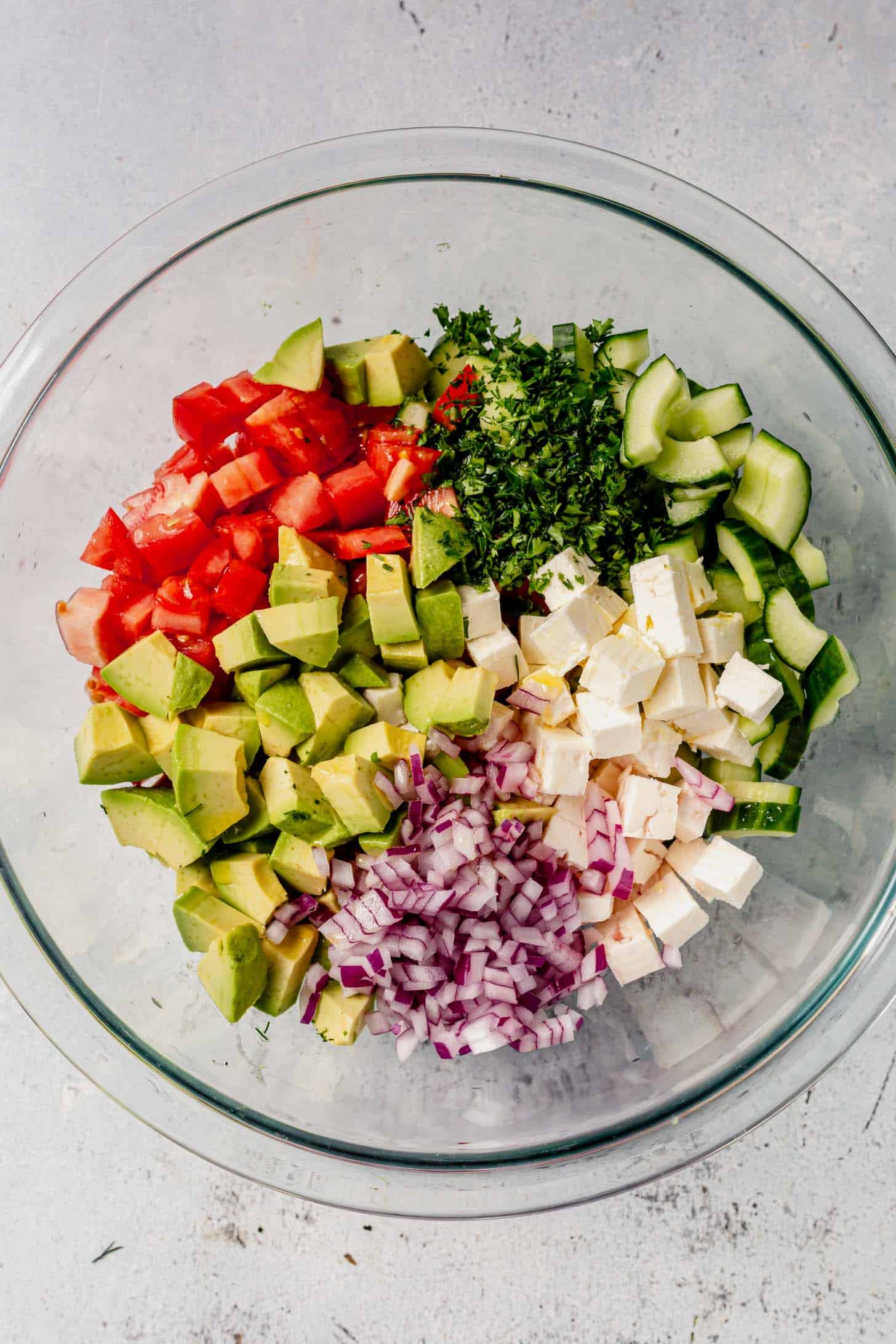 A glass bowl filled with chopped avocado, tomato, cucumber, red onion, feta cheese, and fresh herbs arranged in separate piles beside flaky Greek salmon on a light surface.