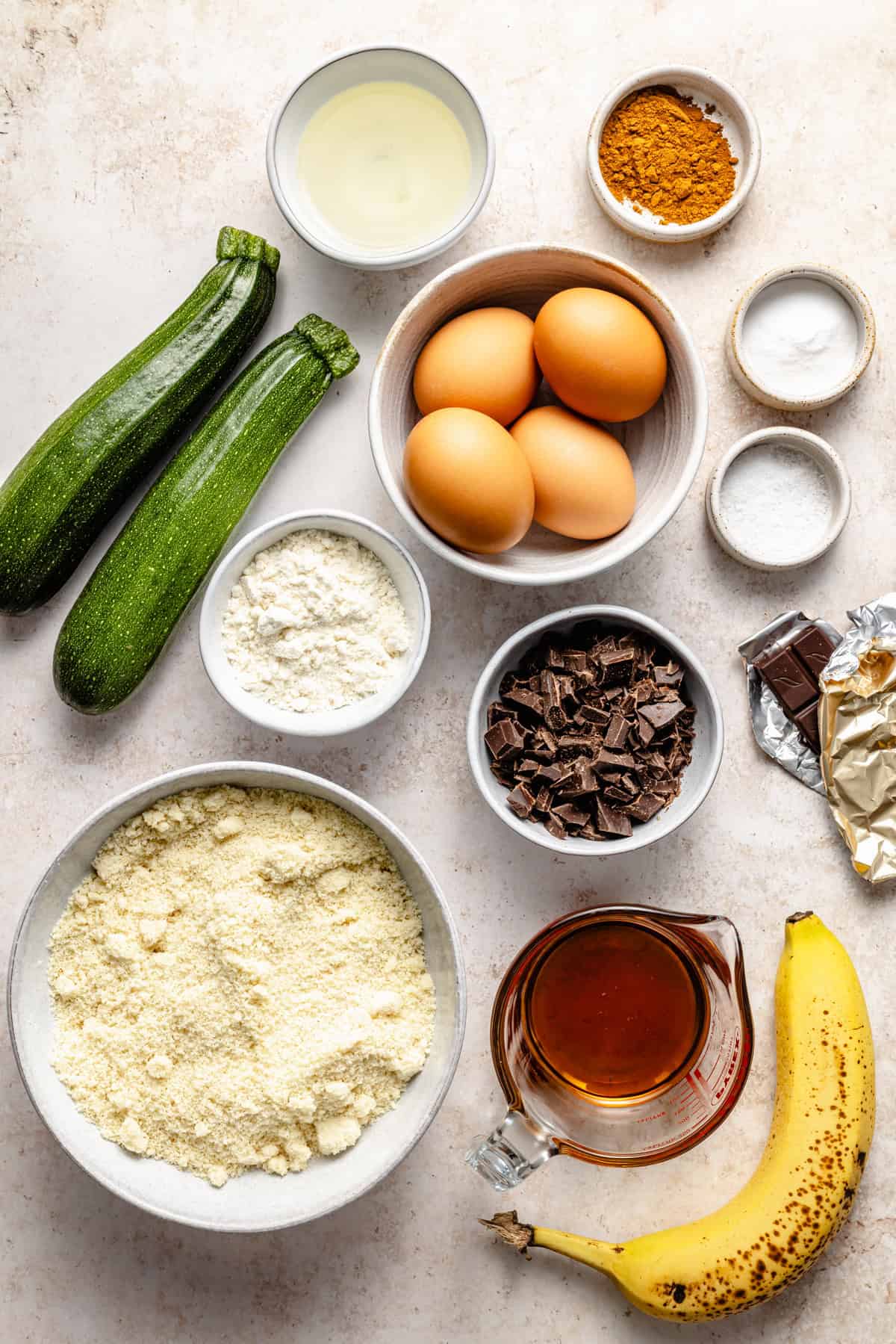 A flat lay of ingredients for paleo zucchini bread on a light surface, including two zucchinis, four eggs, a banana, almond flour, chopped chocolate, maple syrup, oil, baking powder, baking soda, flour, and a bowl of spices.