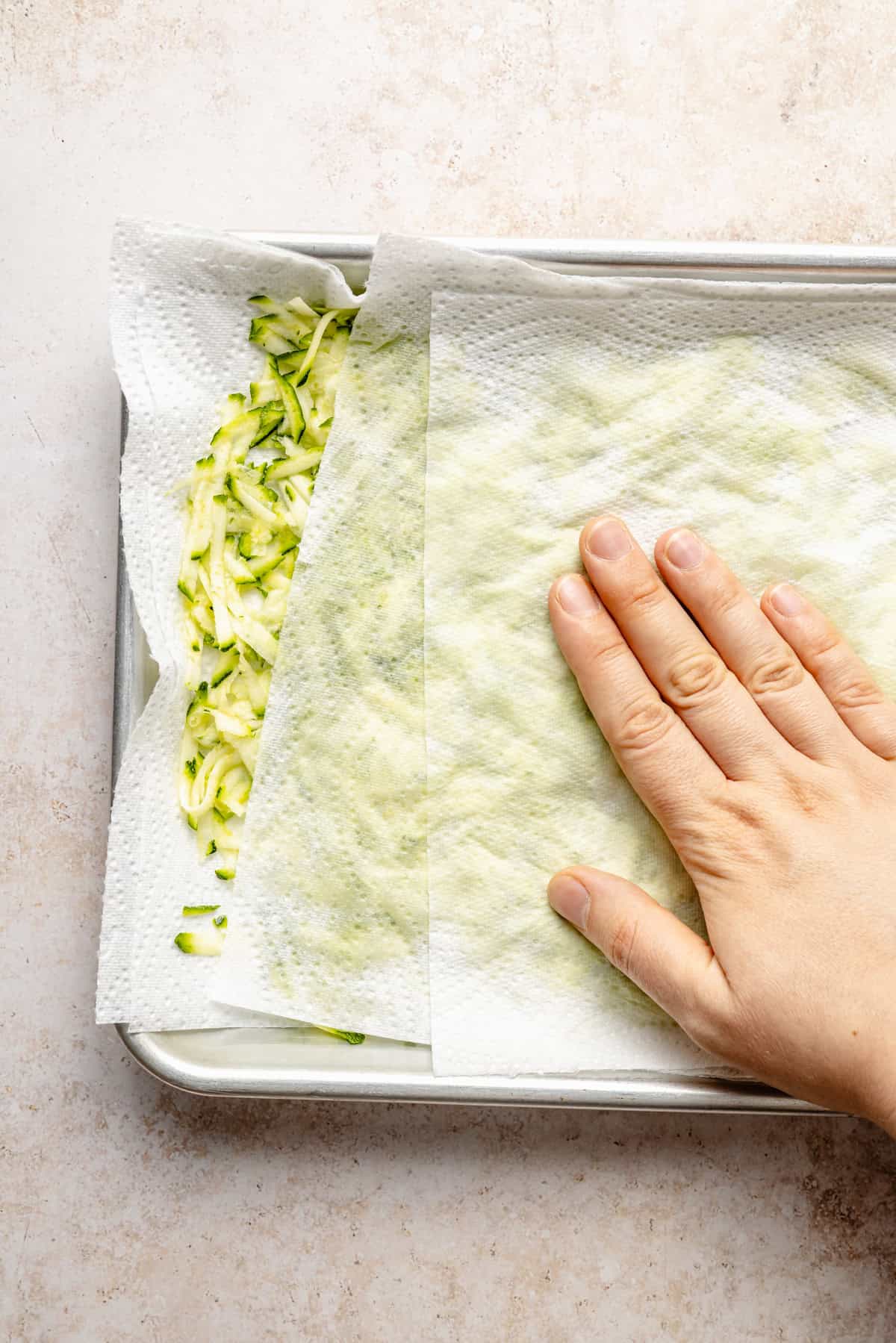 A hand presses a paper towel over shredded zucchini on a baking sheet to absorb moisture—an essential step for making perfectly textured paleo zucchini bread. Another paper towel rests underneath the zucchini on the tray.